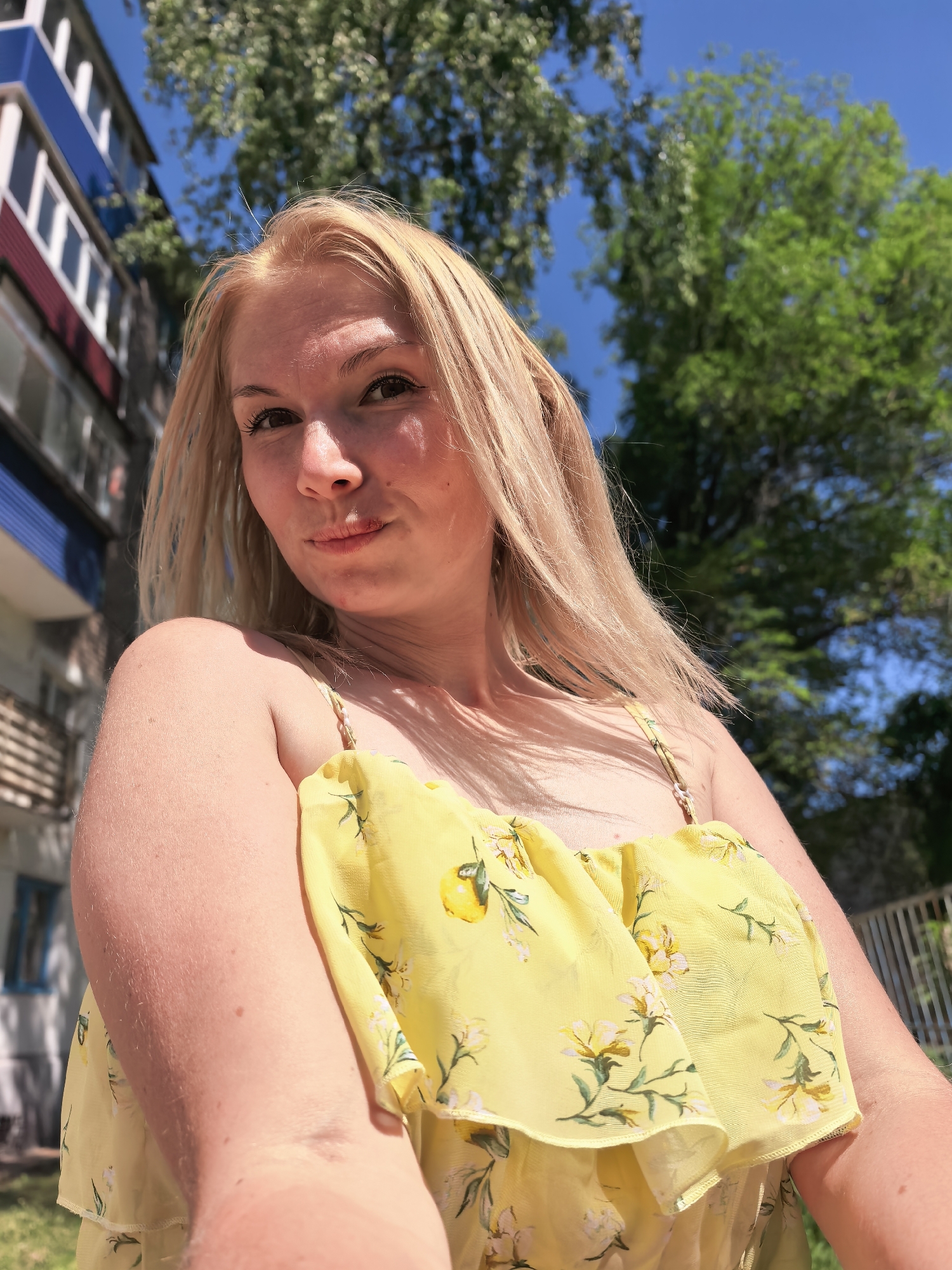 A woman with blond hair is taking a selfie outdoors. She is wearing a yellow top with floral print and small lemon accents. Behind her, there is a blue sky with green trees and a building with a red, white, and blue facade. The location is Sterlitamak, Russia.
