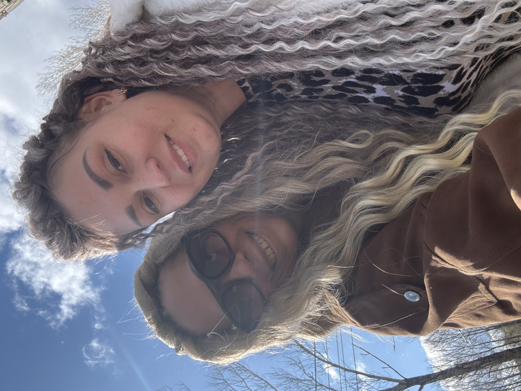 The image is a low-angle selfie featuring two women looking down towards the camera, captured outdoors on a bright, sunny day. The clear blue sky with scattered white clouds forms the background.
The woman on the left (upper part of the original image) has long, voluminous, curly hair with a striking blend of dark and light, possibly grey or blonde, tones. She is wearing a leopard print garment under what appears to be a light-colored, possibly faux fur, collar. An earring is visible in her left ear, and she offers a gentle, open-mouthed smile towards the camera.
The woman on the right (lower part of the original image) has long, wavy blonde hair, which is partly covered by the fur-lined hood of a brown coat or jacket. She is wearing dark sunglasses and also has a cheerful, open-mouthed smile, revealing her teeth.
The photograph is taken from a perspective looking upwards, with bare tree branches visible against the bright sky in the lower right portion of the frame, suggesting a late winter or early spring setting. The bright sunlight creates a warm glow, with some lens flare noticeable on the right side of the image. Both women appear happy and relaxed, enjoying the outdoor moment. The scene takes place in what appears to be a natural or semi-natural outdoor environment, consistent with the given location context of Заволжье, Russia. There is no visible text in the image.