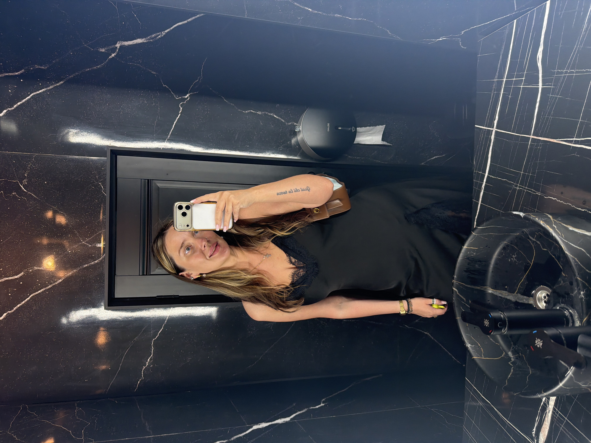 A woman with long, wavy, light brown hair takes a selfie in a black marble-tiled restroom. She is wearing a black slip dress with lace trim, a gold watch on her left wrist, and several bracelets. A small brown crossbody bag is slung over her right shoulder. The woman holds a white smartphone in her right hand, capturing her reflection in a large mirror. The restroom features a black marble countertop with a black sink and faucet. A black paper towel dispenser is mounted on the wall to the right of the woman. The lighting in the room appears to be warm and ambient, reflecting off the marble surfaces.