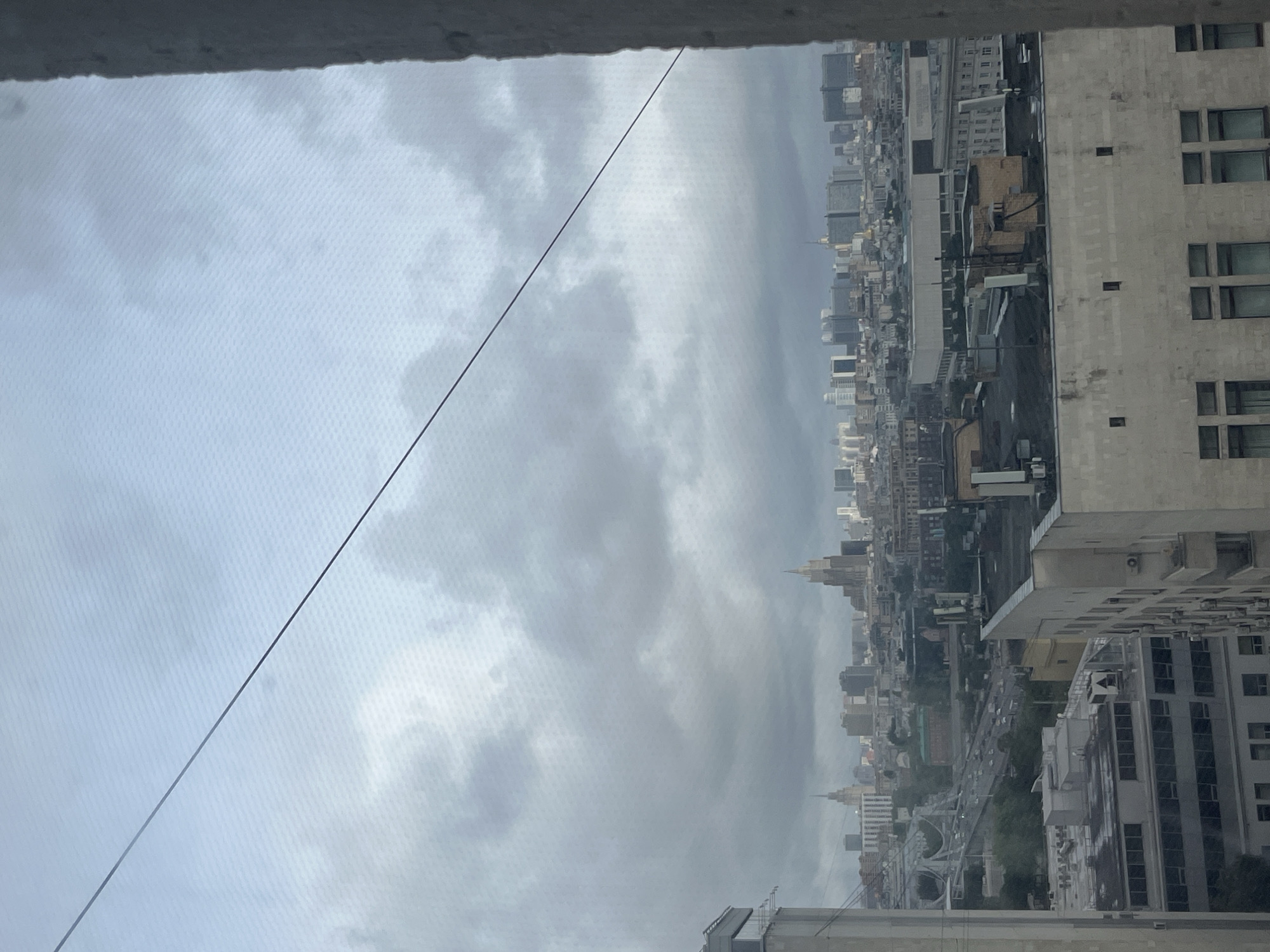 The image shows a cityscape on a cloudy day. The view is from a high vantage point, possibly a window or a balcony. The buildings are dense, varying in height, and are typical of an urban environment. Some buildings have a beige or light gray facade with visible windows. There is a road with traffic visible in the distance, suggesting a busy area. The overall tone of the image is muted due to the overcast sky and the grayness of the buildings. Given the spires in the image, the location of this photo appears to be Moscow, Russia.