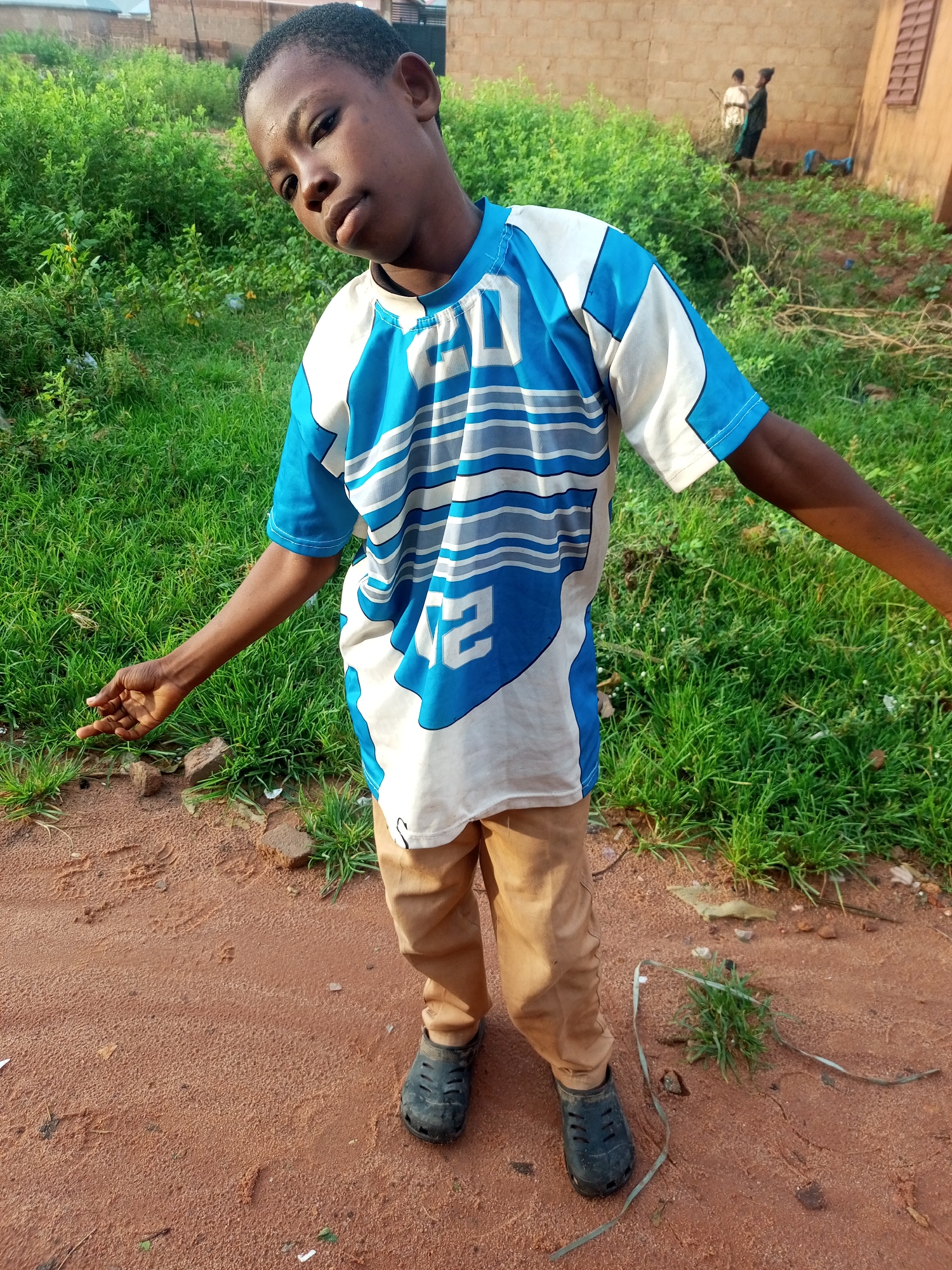 In a dusty, outdoor environment, a young Black male stands facing forward, looking towards the camera with a slight head tilt. He is wearing a blue and white t-shirt with a graphic design and tan pants. His left arm is extended slightly outward with his hand open, palm up, in a gesture. Black rubber shoes are on his feet. The ground beneath him is reddish-brown dirt with some scattered debris, including small stones and dry leaves. Behind him, a patch of lush green grass grows, and further back, a brick building is visible. In the background, two other figures, one in a white shirt and another in a dark outfit, are interacting near the building. The overall setting suggests a casual outdoor scene in a residential area, possibly in Tempure, Nigeria.