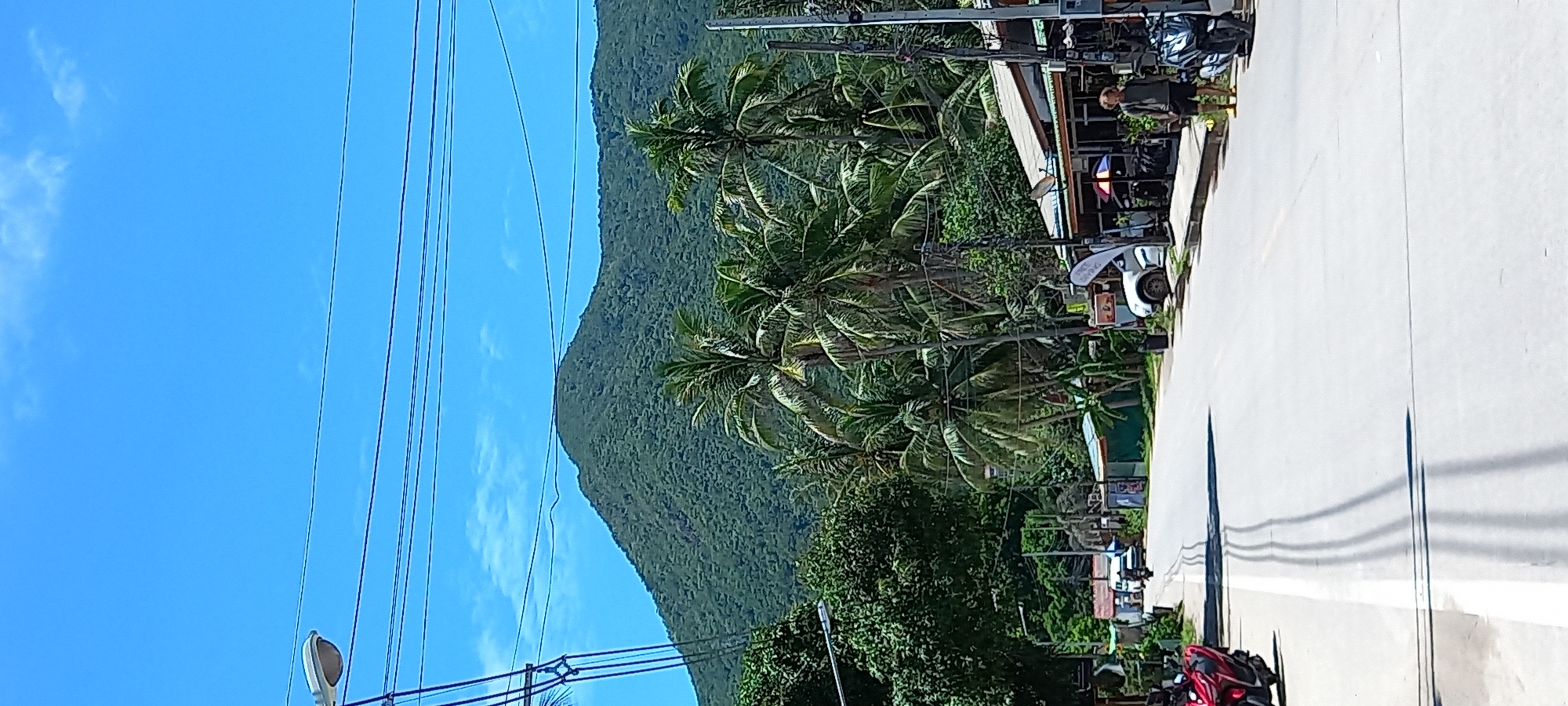 The image depicts a street scene in Baan Chaloklum, Thailand, with a mountain covered in dense green foliage rising in the background and a clear blue sky overhead. Power lines extend across the upper left portion of the frame. Palm trees are interspersed along the roadside. A street with a concrete surface occupies the right portion of the frame. A red scooter is parked on the side of the road. Along the street are buildings with various commercial enterprises. A white vehicle is parked beside one of the buildings, and a person stands in front of the building.