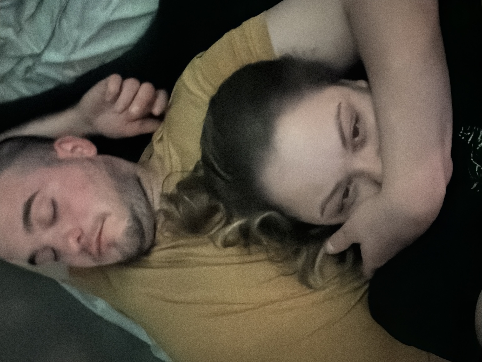 A man and woman are in an intimate embrace, likely in a bedroom. The man lies on his back, eyes closed as if asleep. The woman is positioned on top of him, with her head resting on his chest and arm. Her eyes are open, and she is looking at the camera. They are lying on a yellow shirt on top of a bed with white and black bedding. There is no visual information to determine the location as Varna, Bulgaria.