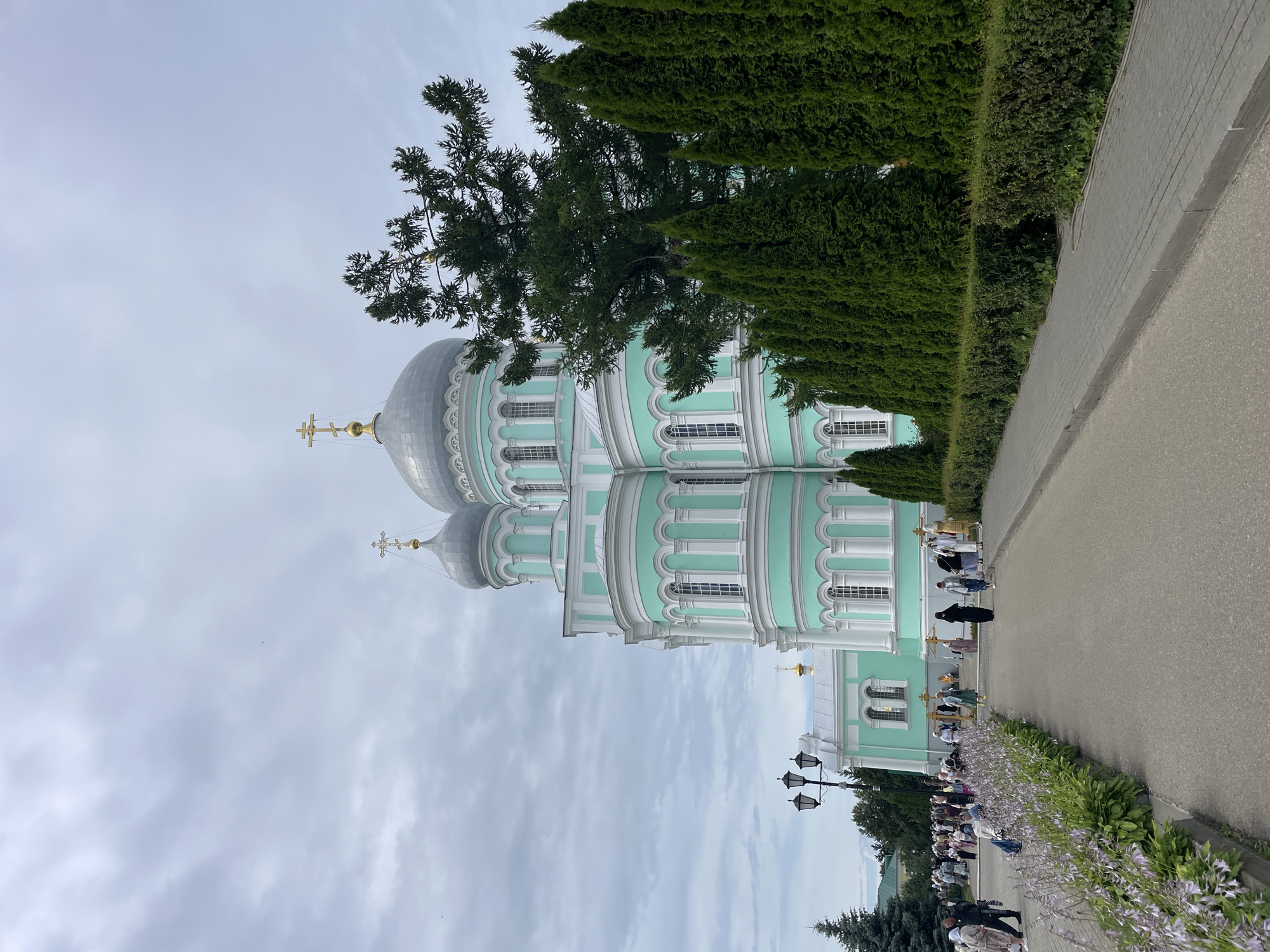 This is a ground-level, vertical, eye-level shot of the Holy Trinity Cathedral in Diveyevo, Russia, on an overcast day. The cathedral is a prominent, large, light turquoise structure with white trim and silver domes topped with golden crosses. It features multiple arched windows and decorative columns. To the right of the cathedral, a row of tall, dark green cypress trees lines a manicured hedge. In the foreground, a paved walkway curves to the right, bordered on the left by a flower bed with light purple blooms and dense green foliage. A paved path leads away from the viewer toward the cathedral. People, dressed in modest attire, are walking along the path and gathered in front of the cathedral. A lamppost with three lights stands to the left of the path. The overall atmosphere is serene and spiritual.