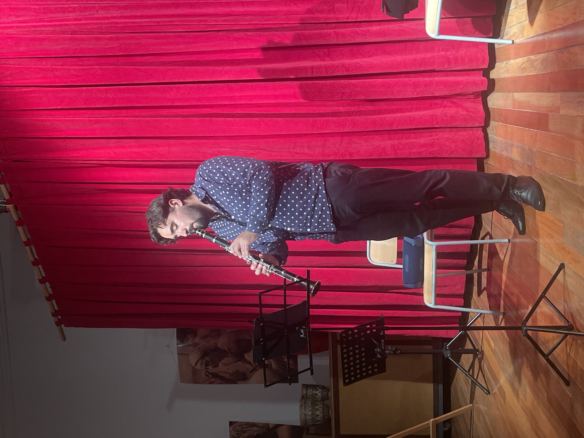 A man is playing a clarinet on a stage. He is wearing a patterned blue shirt and black pants. Behind him is a red velvet curtain. There are two music stands in front of him, and a chair is to his right. The floor is made of wood. On the wall to the left, there are framed images. The image doesn't provide sufficient information to identify the specific location in Barcelona.