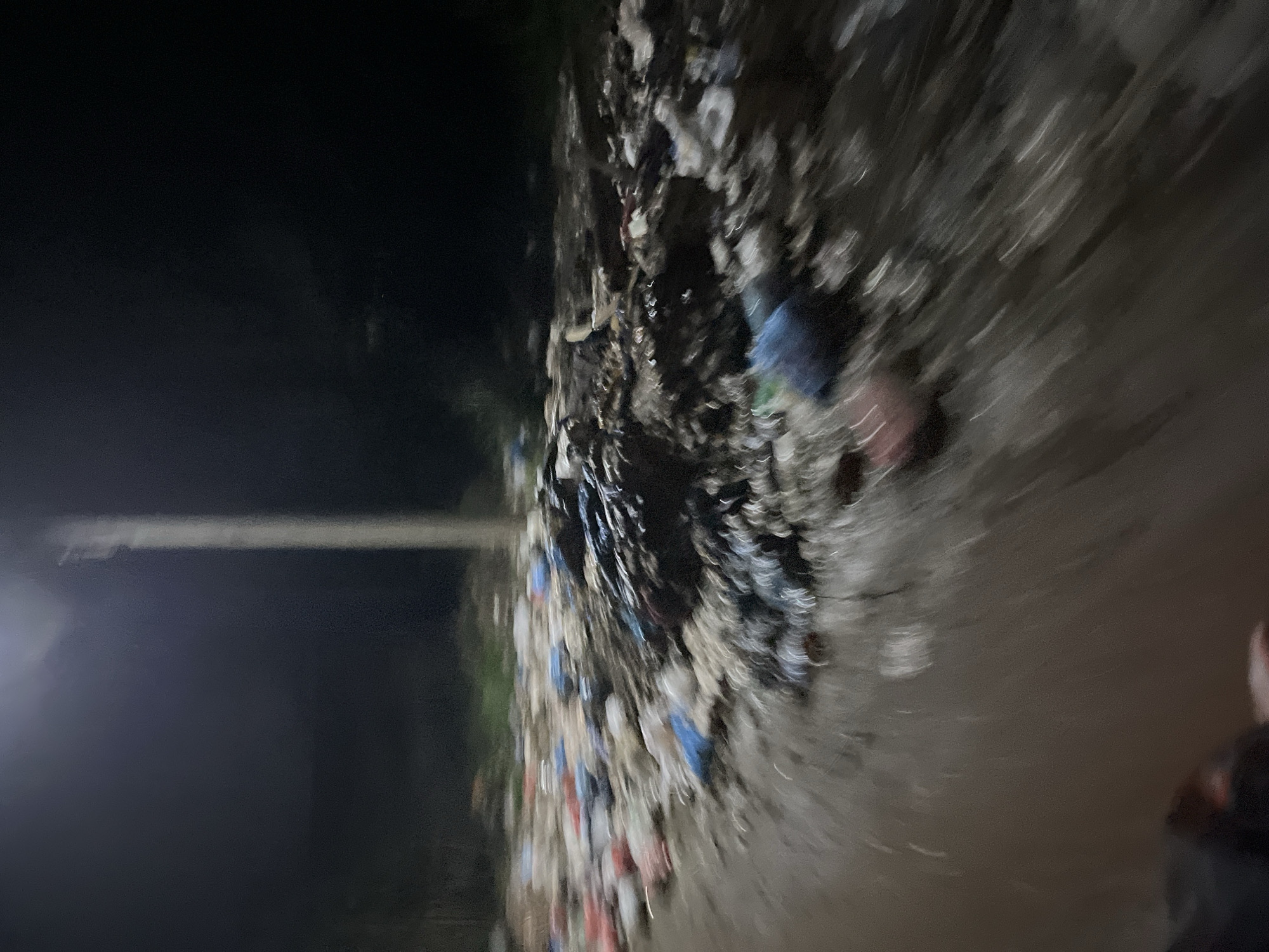 The image depicts an outdoor scene at night, illuminated by a single, tall light pole casting a bright beam downwards. The foreground is dominated by a large pile of refuse, consisting of various types of trash, including plastic bags, containers, and debris. The ground appears to be wet or muddy, with water or liquid present around the garbage. The overall environment is dark and appears to be an open area, possibly on the outskirts of a city or in a less developed part of Benuaraja, Indonesia, given the context. The image quality is poor due to low light and motion blur, making it difficult to discern fine details of the trash or any potential inhabitants of the scene. There are no discernible people actively engaged in any action within the frame.