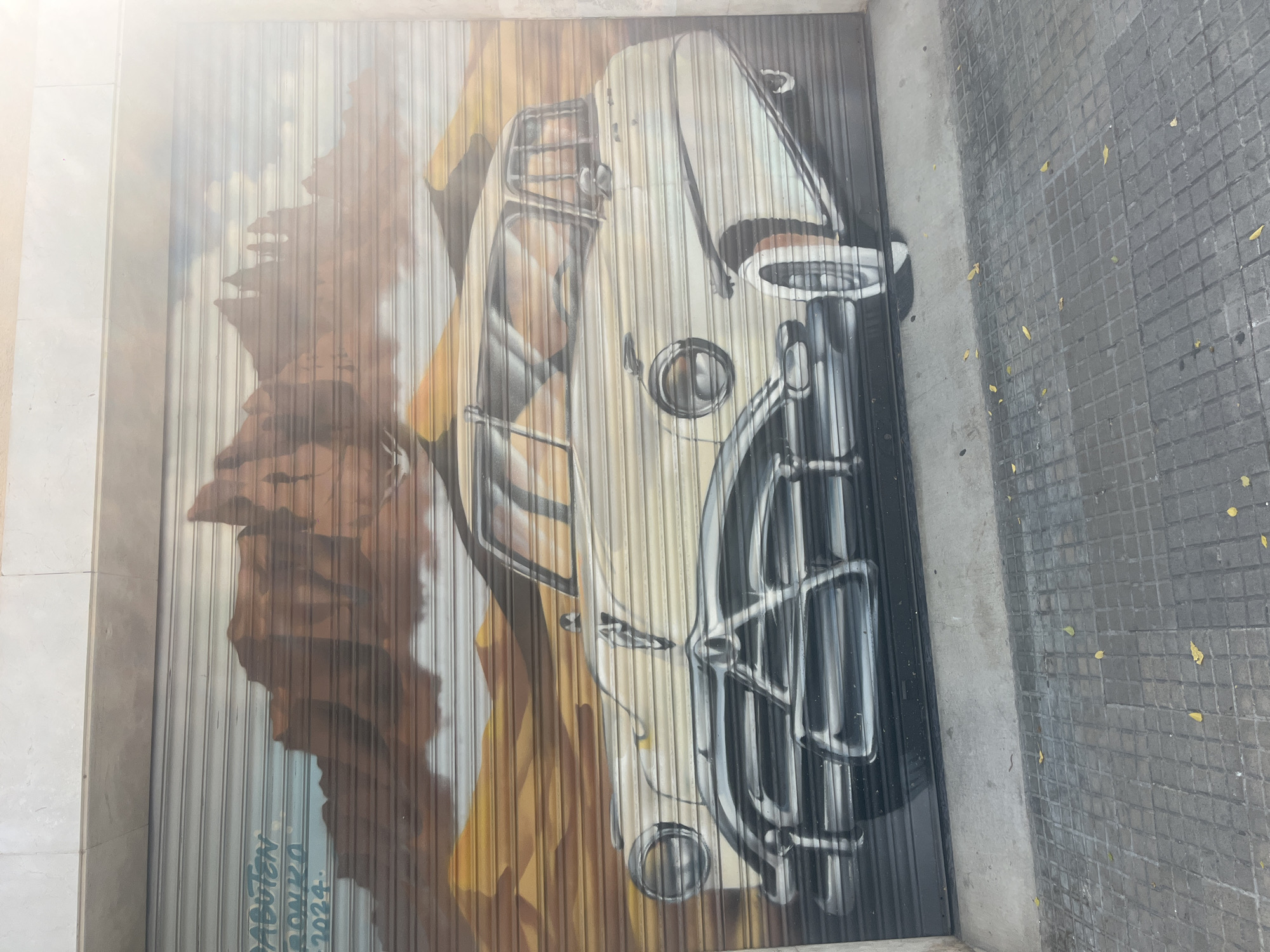 The image shows a mural painted on a corrugated metal security shutter. The mural depicts a vintage cream-colored car parked in a desert landscape with reddish-brown mountains in the background and sand dunes in the foreground. The car is a classic model with chrome accents on the grille, headlights, and trim. The sky above is a pale blue with wispy white clouds. In the upper left corner of the mural, text is visible in light blue and green paint, appearing to read "DADUBTEN BONKA 2024".
The security shutter is located on the facade of a building, with a light-colored concrete wall above it. Below the shutter, a sidewalk paved with square gray tiles is visible. A few yellow leaves are scattered on the sidewalk. The scene is illuminated by a bright light, possibly sunlight, which causes a lens flare in the upper right portion of the image. The location is identified as Tarragona, Spain.