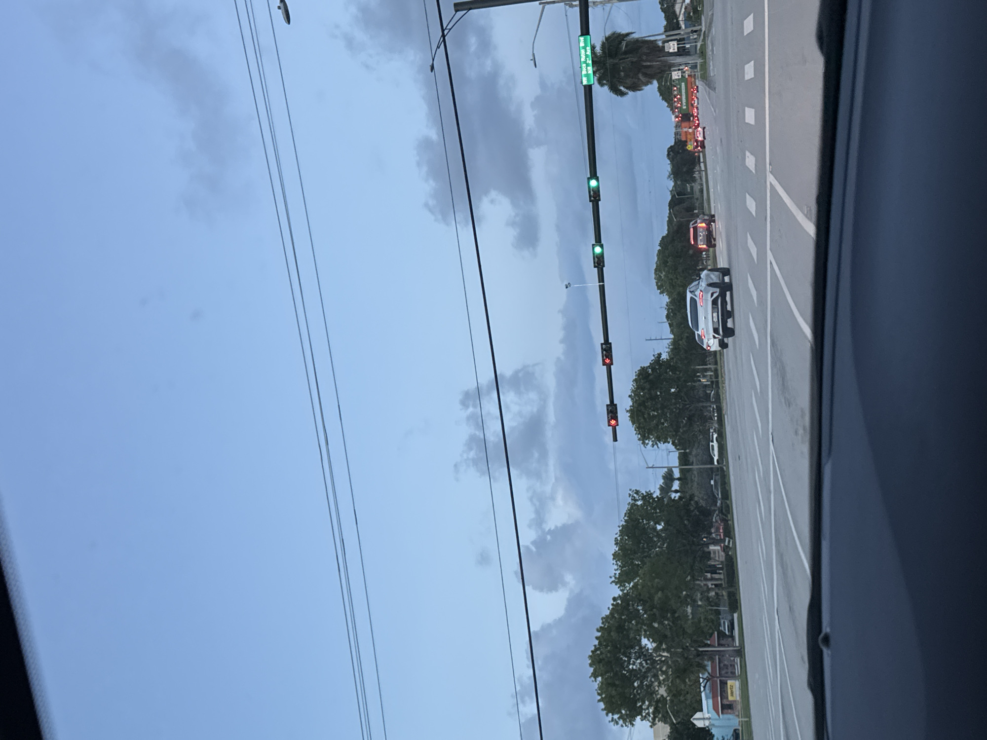 The image shows a view from inside a vehicle looking out onto a street in Boca Raton, Florida. The sky is overcast with gray clouds. There are multiple utility lines running across the top of the frame. A traffic light is visible, with a street sign indicating "NW Boca Raton Blvd" and "NW 2nd Ave". The traffic lights are showing green signals in some locations and red signals in others. A white SUV is on the right side of the road, moving in the same direction as the vehicle taking the photo. There is a marked crosswalk. Trees line the side of the road. In the distance, there appears to be traffic congestion.