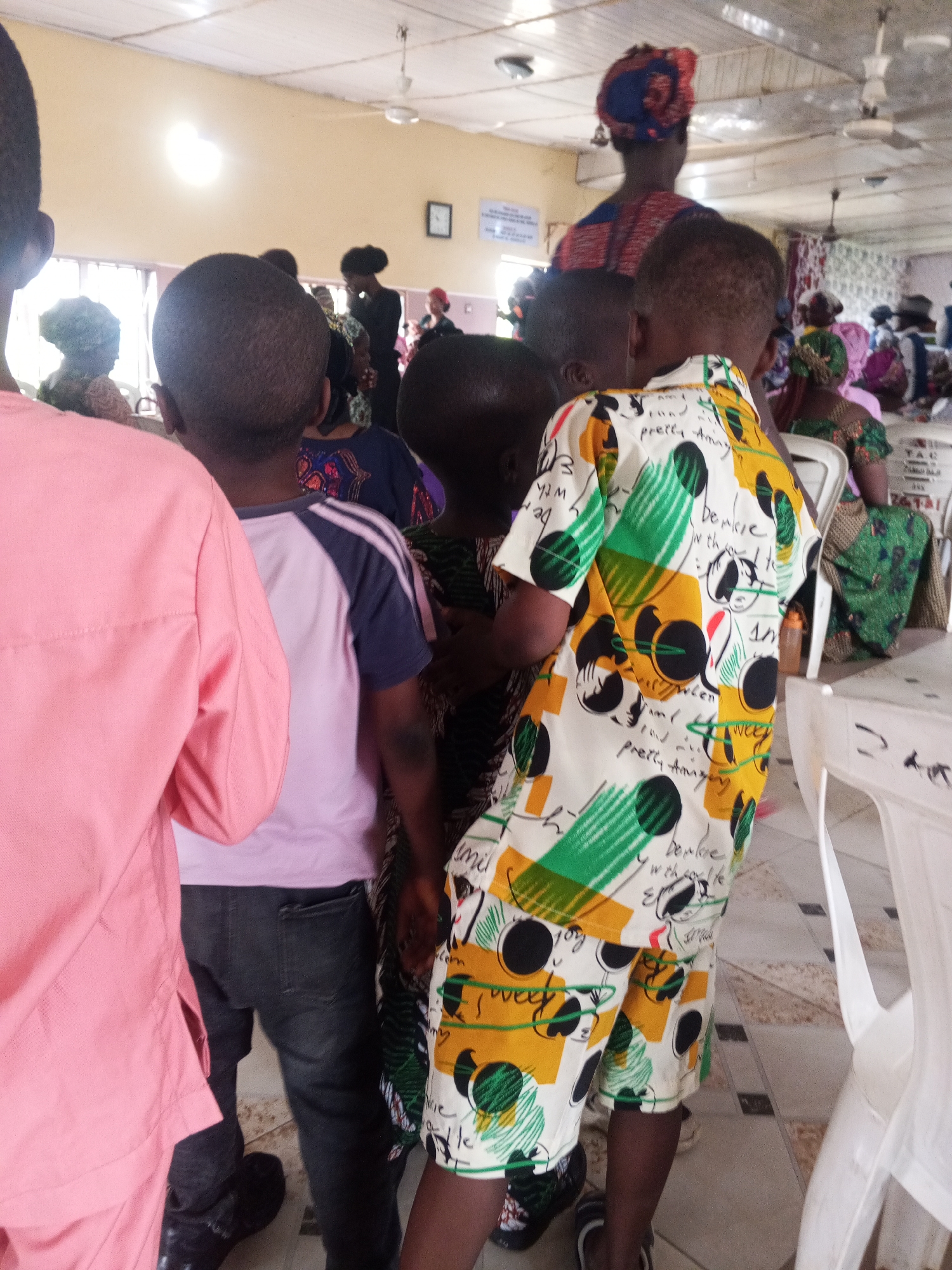 In the city of Zango-ndaji, Nigeria, a group of people are gathered indoors. The scene appears to be a community event or a religious service. In the foreground, a young boy is wearing a colorful, patterned short-sleeved shirt and shorts. To his left, another young boy is wearing a pink long-sleeved shirt, and behind him, a person in a purple shirt with dark sleeves. Further back, several women are visible, some wearing headwraps and patterned clothing. The room has a tiled floor, a white ceiling with a fan, and fluorescent lighting. A noticeboard with text is mounted on the wall. White plastic chairs are arranged in rows, suggesting a seating arrangement for attendees. The overall atmosphere seems to be one of a lively gathering.