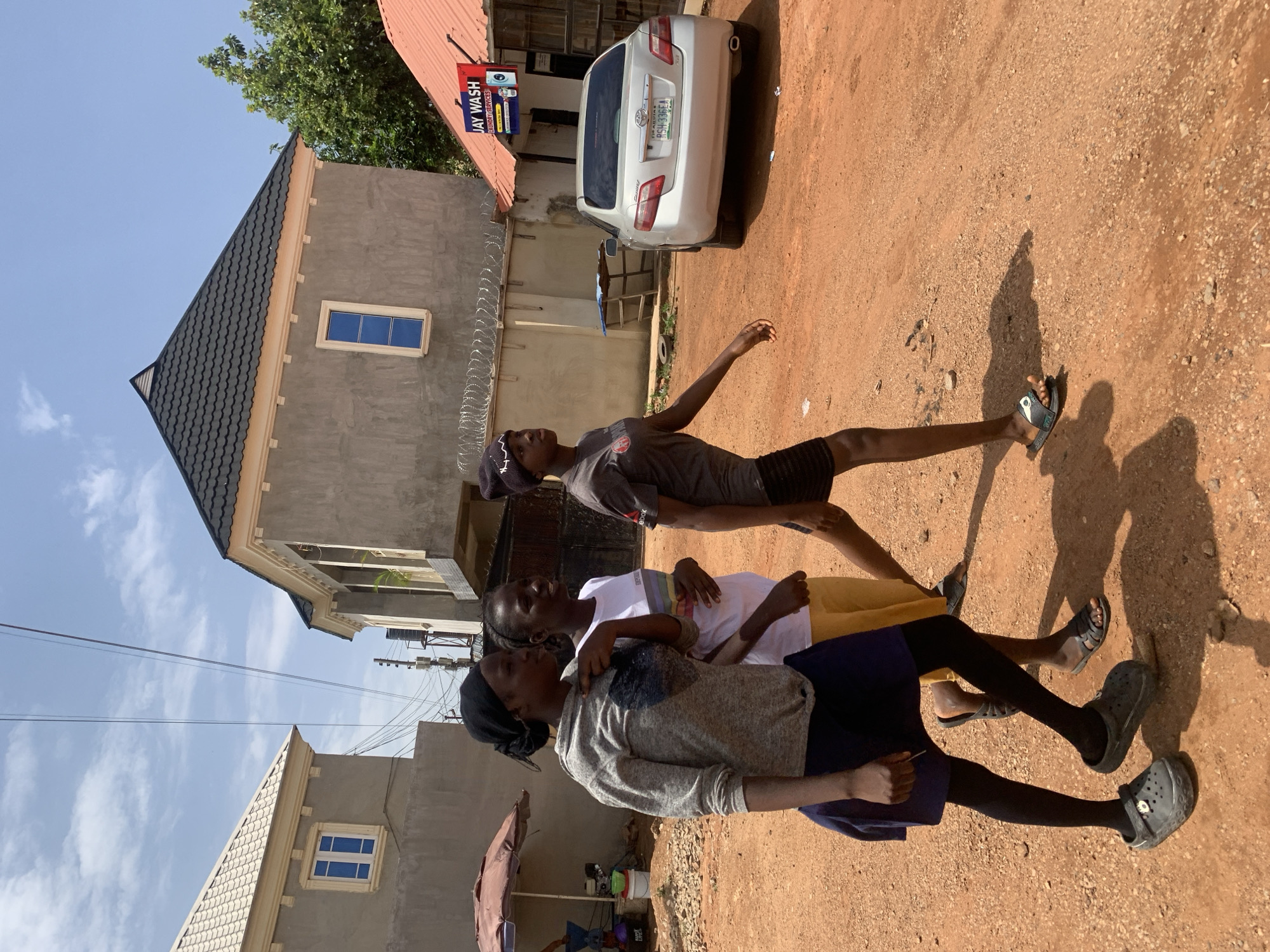 Three people stand on a dirt road. One person wears a grey sweater and a navy blue skirt. Another person wears a white shirt and a yellow skirt. The third person wears a grey t-shirt and black shorts. The background shows buildings, a car with license plate RSH-336EJA, a sign for "Jay Wash", and a blue sky with clouds. This scene appears to be located in Gura Top, Nigeria.