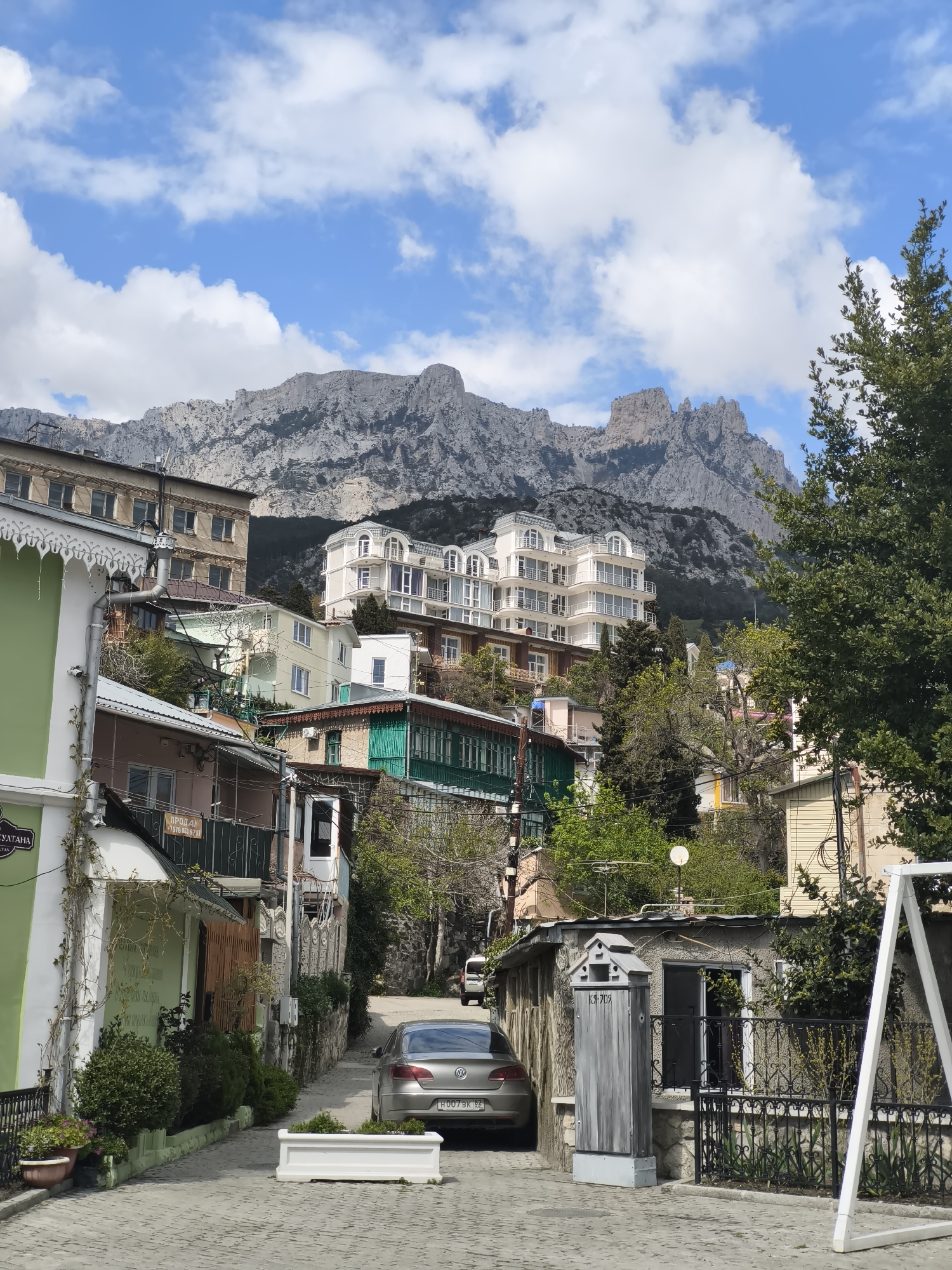 The image shows a street scene in the city of Алупкинский городской совет, Ukraine. A cobblestone street slopes uphill, lined with buildings on either side. A silver Volkswagen sedan is parked on the right side of the road, its license plate partially visible. A small gray building with a black wrought iron fence stands adjacent to the parked car, along with a white swing frame. The buildings on the left side of the street are varied in color and style, with some featuring vines. In the background, a large, rocky mountain rises above the buildings, with a partly cloudy sky. Trees and vegetation are interspersed among the buildings.