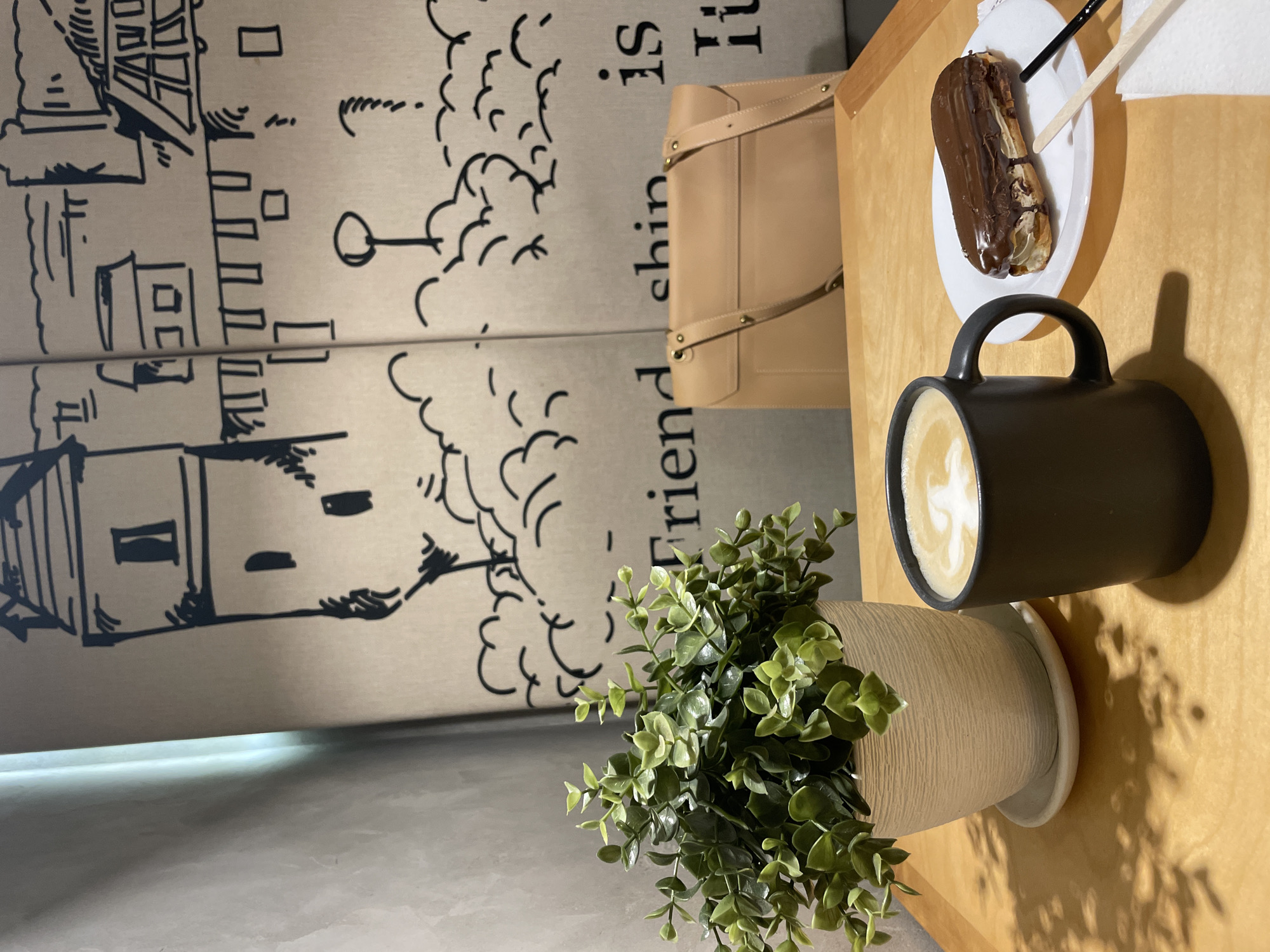 The image shows a close-up of a table setting, likely in a cafe in Krasnoyarsk, Russia, based on the common style and items present. The table is light-colored wood and features a black mug filled with a latte or cappuccino, with latte art visible in the foam. Beside the mug is a white plate with a chocolate éclair. There are also two sticks, possibly for stirring the coffee or eating the éclair. Next to the coffee is a small potted plant with green leaves. In the background, there is a beige handbag and a decorative wall panel with a drawing of a city scene and the word "Friendship" in Russian. The overall lighting is bright, casting shadows from the objects onto the table.