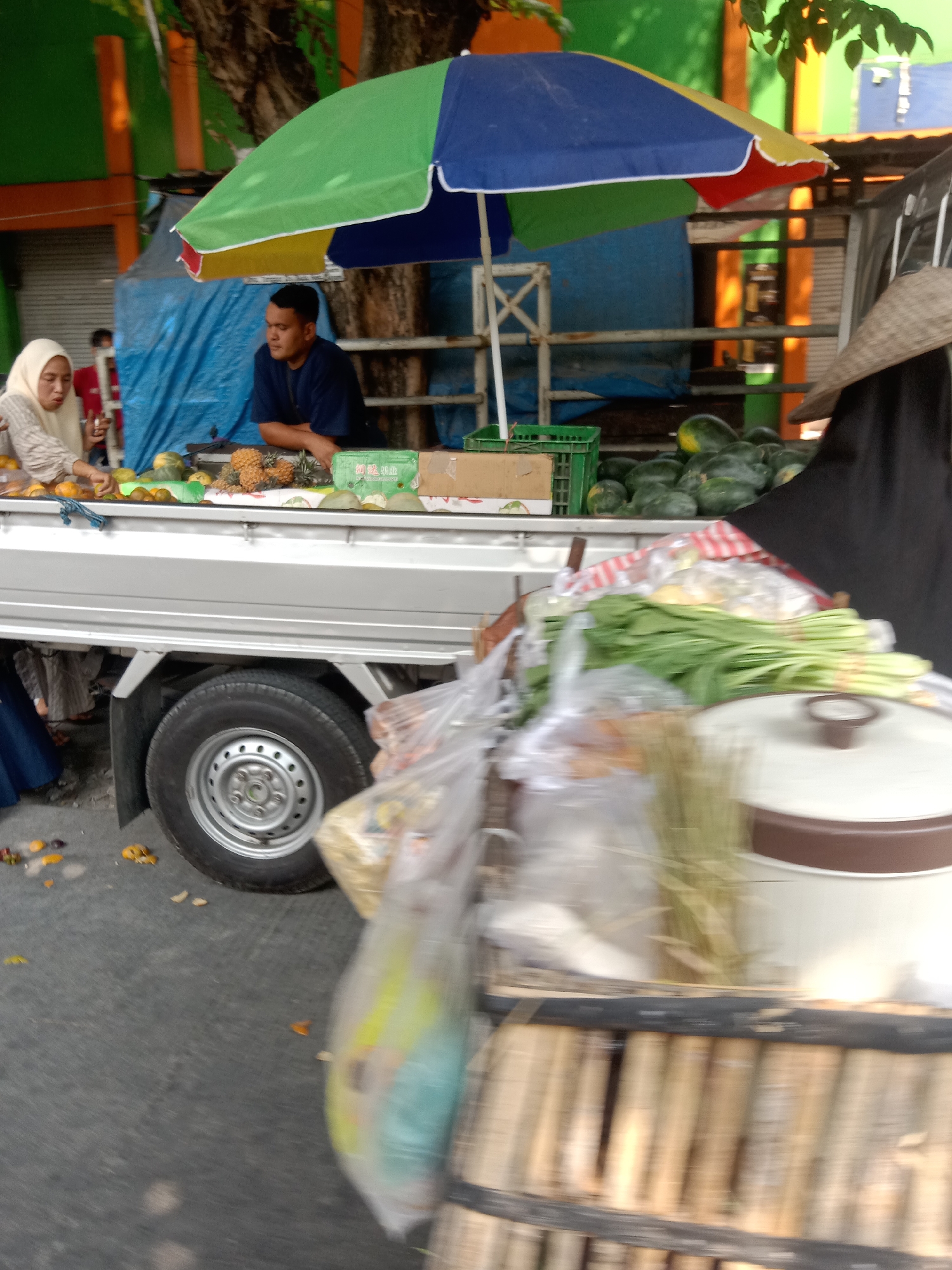 A low-angle, slightly blurred outdoor shot captures a fruit stand in Sugihwaras, Indonesia. A colorful umbrella, segmented in green, blue, yellow, and red, is tilted to the right, providing shade. Two individuals are partially visible. On the left, a woman wearing a light-colored hijab and patterned shirt is seen from the chest up, tending to fruit on a silver truck bed. Her hands are busy arranging or inspecting the produce. To her right, a man in a dark blue shirt is seated behind a display of pineapples and other fruits, looking to his left. The truck bed is laden with various fruits, including pineapples, melons, and what appear to be watermelons. In the background, a blue tarp is visible behind the umbrella, and a tree with textured bark looms over the scene. To the right of the frame, a person in a black abaya and conical hat stands partially obscured, facing away from the camera. The foreground is dominated by a close-up of a cart filled with leafy green vegetables and a large white pot with a brown lid. The street pavement is visible below, with scattered leaves and fruit rinds.