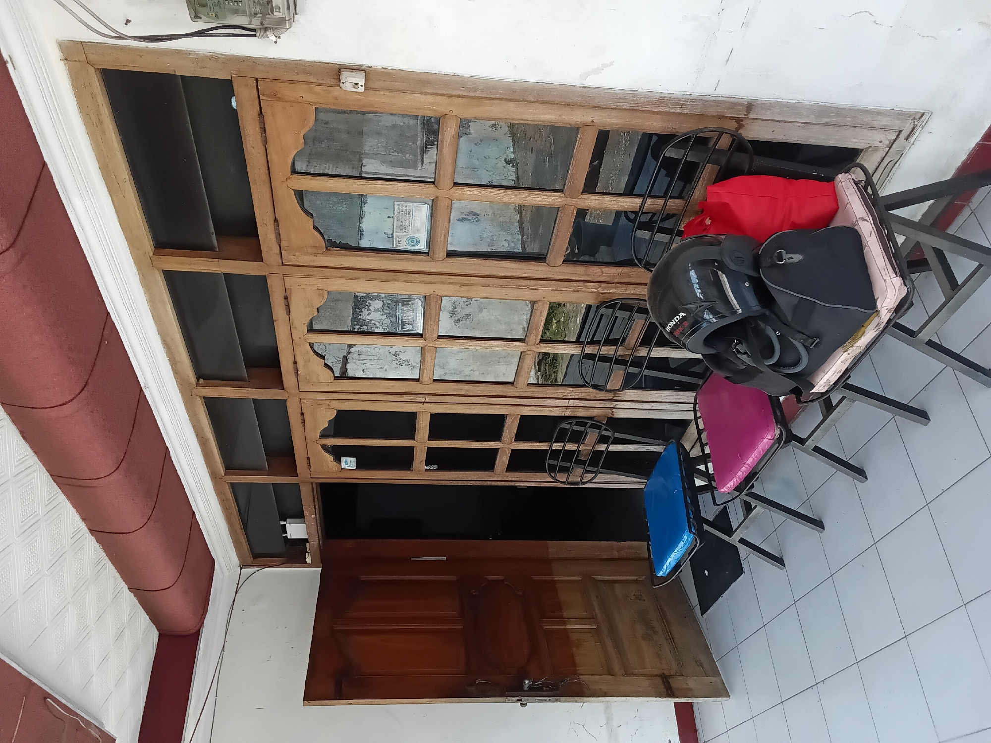 A house entrance in Kelutan, Indonesia features a wooden door opened to reveal an interior hallway. In front of the entrance are two metal folding chairs. One chair has a blue cushion, and the other has a pink cushion. On the pink cushion rests a black motorcycle helmet and a black duffel bag, with a red bag leaning against the helmet. The house has a wooden window frame with multiple panes and a white wall. A pipe covered in red fabric runs along the ceiling. An electrical meter is mounted on the exterior wall to the right of the entrance. The ground is tiled with white square tiles.