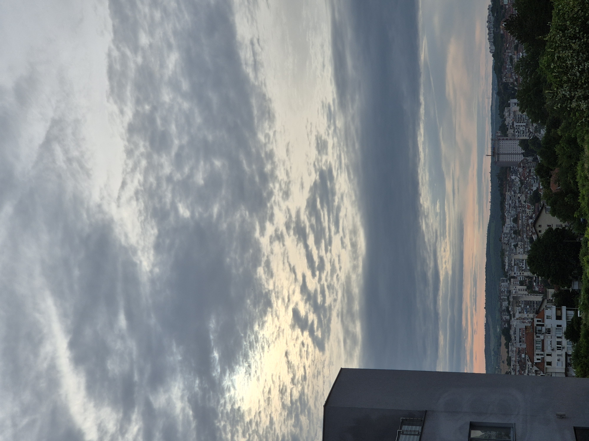 An exterior, high-angle, full shot captures a cityscape under a cloudy sky. The left side of the frame is dominated by the corner of a modern, grey, multi-story building with a balcony. Beyond this building, a dense urban landscape unfolds, characterized by a variety of buildings, from smaller, older residential structures with tiled roofs to taller, modern apartment blocks and a prominent, tall, light-colored building in the distance. Lush green trees and foliage line the foreground and middle ground of the city, interspersed among the buildings. The sky is filled with a textured pattern of grey and white clouds, with a brighter, more illuminated area suggesting the presence of the sun behind the cloud cover. The overall lighting suggests either early morning or late evening. The location is identified as Unknown, Unknown.