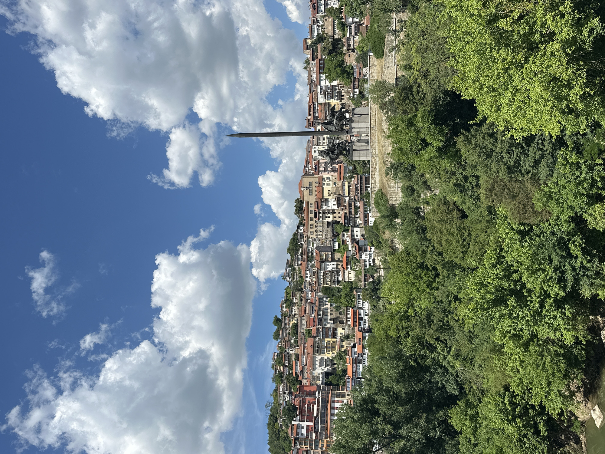 The image depicts the historic city of Veliko Tarnovo, Bulgaria, featuring a panoramic view of its densely packed houses cascading down a hillside under a bright blue sky dotted with fluffy white clouds. In the mid-ground, a prominent monument stands on a raised platform, characterized by a tall, slender obelisk and a sculptural group depicting figures on horseback. Lush green trees form a dense foreground and also dot the hillsides, contrasting with the terracotta roofs and colorful facades of the buildings. The architecture is predominantly traditional, with a mix of stone and painted structures, suggesting a rich historical heritage. The overall scene captures a picturesque, sunlit day in the city, highlighting its unique topography and architectural charm.