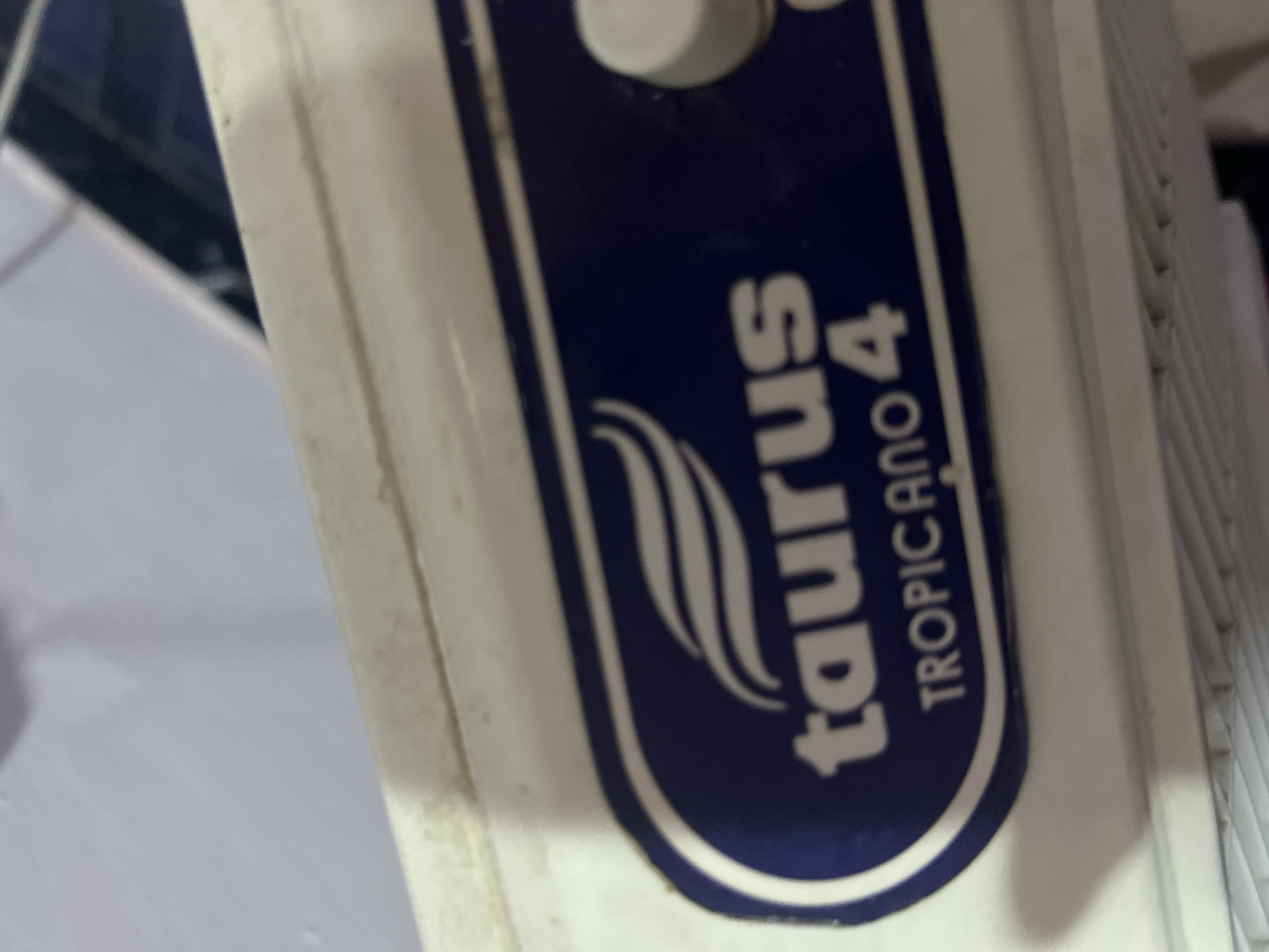 The image shows a close-up of a white Taurus Tropicana 4 electric fan. The model name is printed in white on a blue, rounded rectangle label with a logo that looks like stylized air currents. A white button is visible above the label. The location is not discernible from the image.
