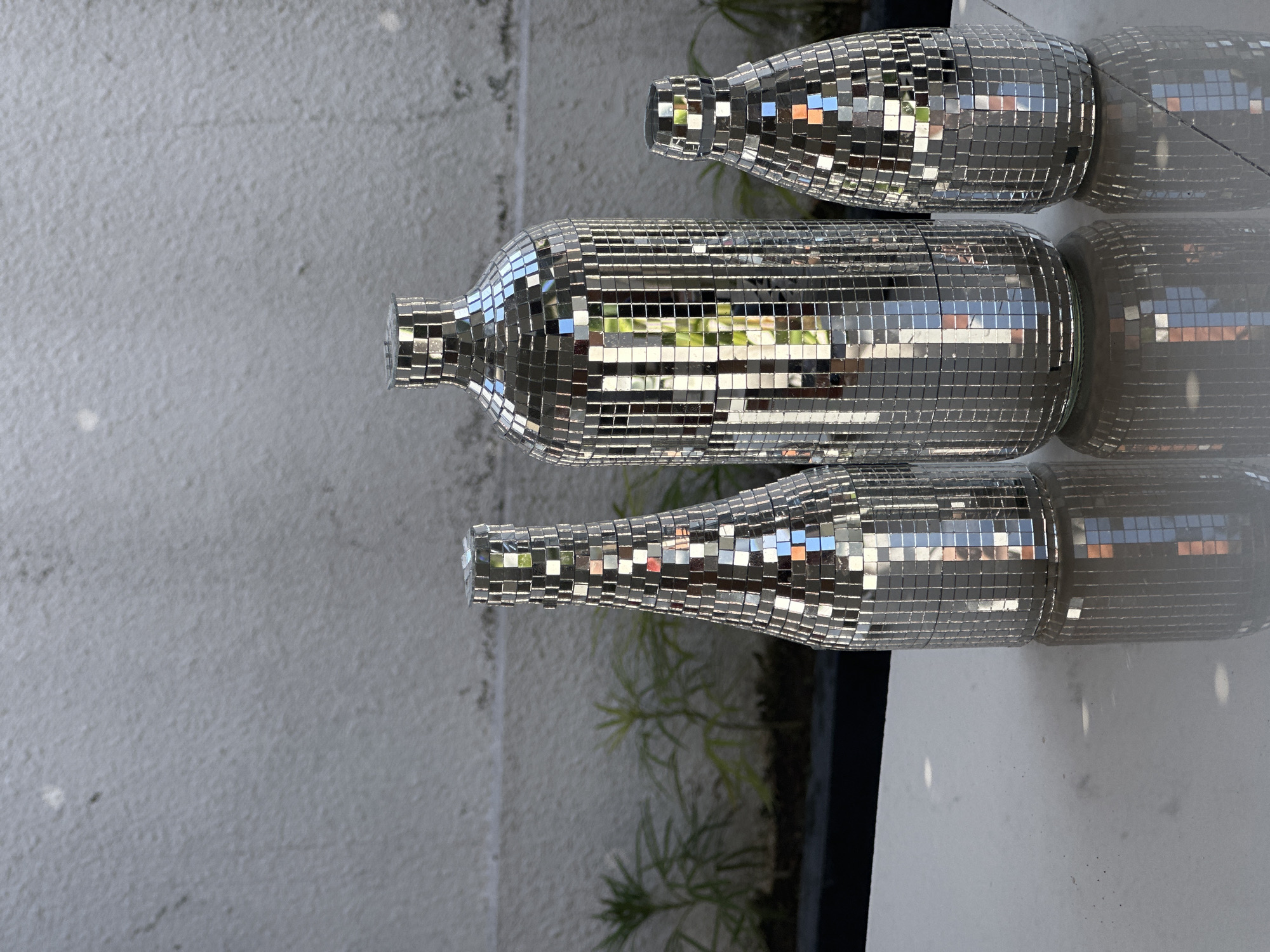 Three disco ball vases of varying heights are arranged on a white tiled surface. The tallest vase is in the center, with a shorter, wider vase to its right, and the shortest, narrowest vase to its left. The vases are covered in small, square mirrored tiles that reflect the surrounding environment, including greenery and a textured grey wall. The reflection of the vases is visible on the polished white surface. The scene appears to be outdoors, possibly in a courtyard or garden setting in North Kuta, Indonesia, given the hint. There are no people present in the image.