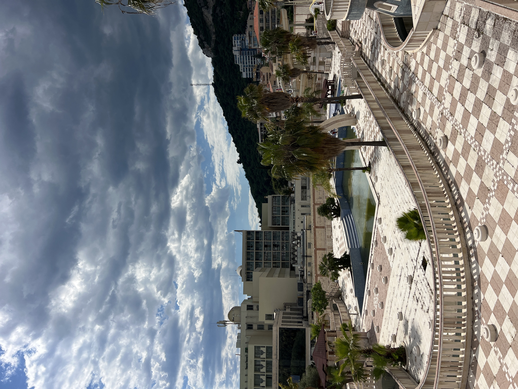 The image captures an elevated view of a section of the Dukley Hotel & Resort in Boreti, Montenegro. Dominating the frame are buildings with light exteriors and many windows, hinting at a luxurious or residential complex. Lush, green palm trees punctuate the scene, providing a tropical ambiance. A decorative, patterned walkway in a checkerboard design stretches into the foreground, flanked by a balustrade. A body of water, likely a pool or decorative pond, runs parallel to the walkway, featuring palm trees at intervals. To the left, a dramatic sky filled with dark, stormy clouds transitions into lighter, partly cloudy areas. In the distance, a mountainous landscape covered in greenery adds depth to the image.