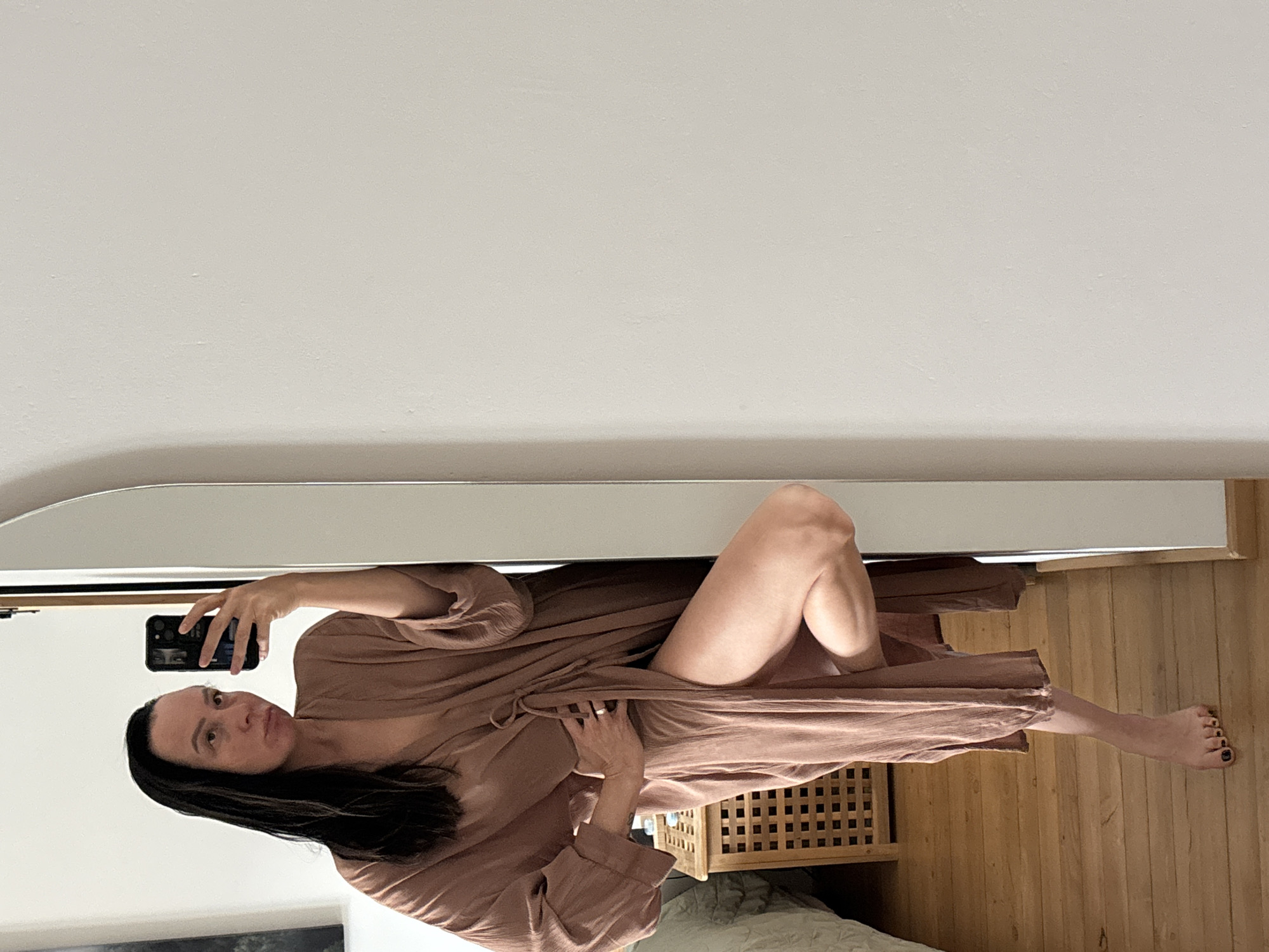 A woman is posing for a selfie in a mirror indoors. She is wearing a brown robe. Her hair is dark. She is holding a smartphone. In the background, there is a white wall, a wooden floor, a wooden storage box, and part of a bed. There is no indication of the location being in the city of Chaikovsky, Russia.