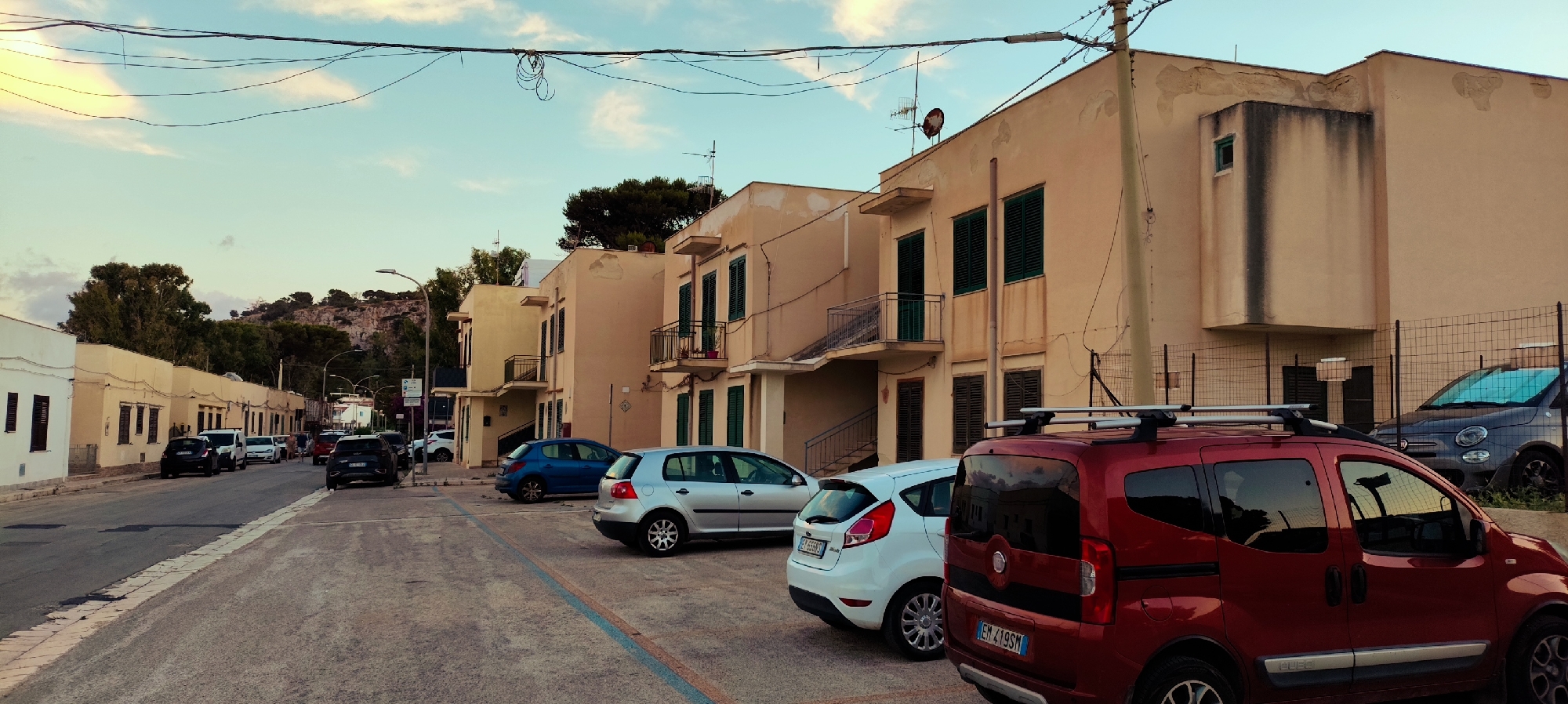 The image depicts a street scene in San Vito Lo Capo, Italy, characterized by modest, two-story buildings with tan stucco exteriors and dark-shuttered windows. The architecture suggests a residential or small commercial area. Several vehicles are parked along the street, including a prominent red van in the foreground, a silver hatchback, a dark blue hatchback, and a dark SUV further down the road. A dark gray Fiat 500 is parked behind a fence on the right side of the image. The street is paved with asphalt and lined with a curb. Overhead power lines are visible against a pale blue sky with scattered clouds. Vegetation, including trees and shrubs, can be seen in the background, with a rocky hillside in the distance. The overall impression is one of a quiet, late afternoon or early evening in a coastal Italian town.