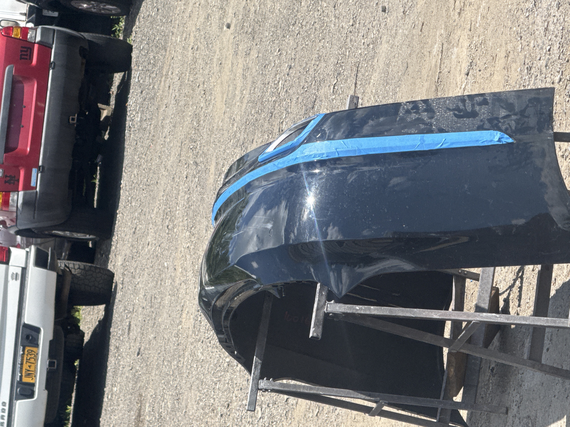 A black car bumper is supported by two metal stands on a gravel lot. The bumper has blue tape on its surface, indicating it is being prepped for repair or painting. In the background, the rear ends of two vehicles are visible: a white pickup truck with license plate JNY-7539 and a red SUV with "NY" decals. The scene appears to be outdoors in an industrial or automotive repair area.