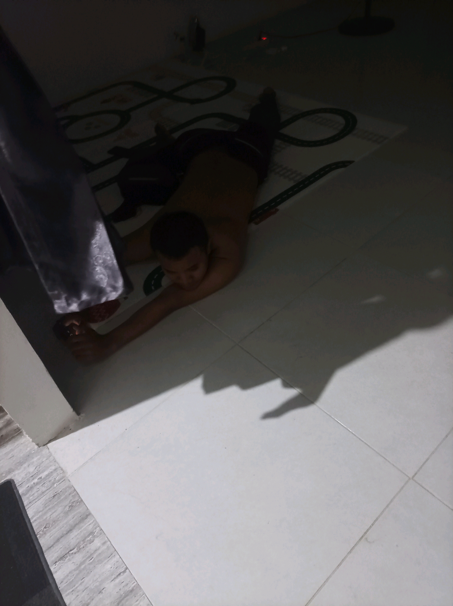 In Canga'an, Indonesia, a person lies on their stomach on a white tiled floor, next to a rug with a car track design. The person is wearing dark shorts and is shirtless, with their left arm extended and hand resting on a red object, possibly a shoe or slipper. Their head is turned to the left, and they appear to be looking at something in their hand. The room is dimly lit, with shadows casting from the person and objects in the scene. A dark grey curtain hangs on the left side of the image, partially obscuring the view. The overall environment suggests a residential indoor space.