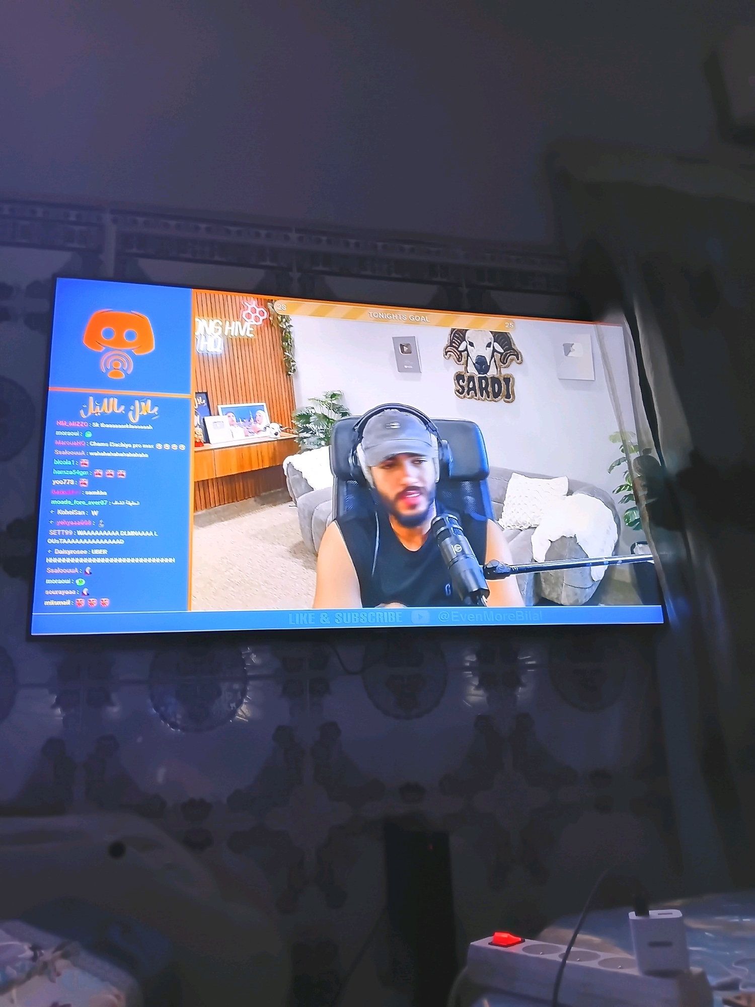 The image depicts a screen displaying a live stream. On the left side of the screen, a blue panel shows the Discord logo and a chat feed with usernames and messages, some of which are in Arabic. The right side of the screen shows a man with a beard and a cap wearing headphones and speaking into a microphone. He is seated in a gaming chair in front of a wall decorated with a ram's head logo and the word "SARDI". In the background, there is a couch and a plant. The bottom of the screen features a banner with the text "LIKE & SUBSCRIBE" and a social media handle "@EvenMoreBilal". The overall setting appears to be a room where a streamer is broadcasting. The presence of Arabic text and the social media handle suggest the stream may be originating from or targeted towards an Arabic-speaking audience. The image is taken from an angle that captures the screen, with some of the surrounding environment visible, including a patterned wallpaper and electrical outlets. The context does not provide specific details to pinpoint the exact location within Guelmim, Morocco, beyond it being a room in the city.