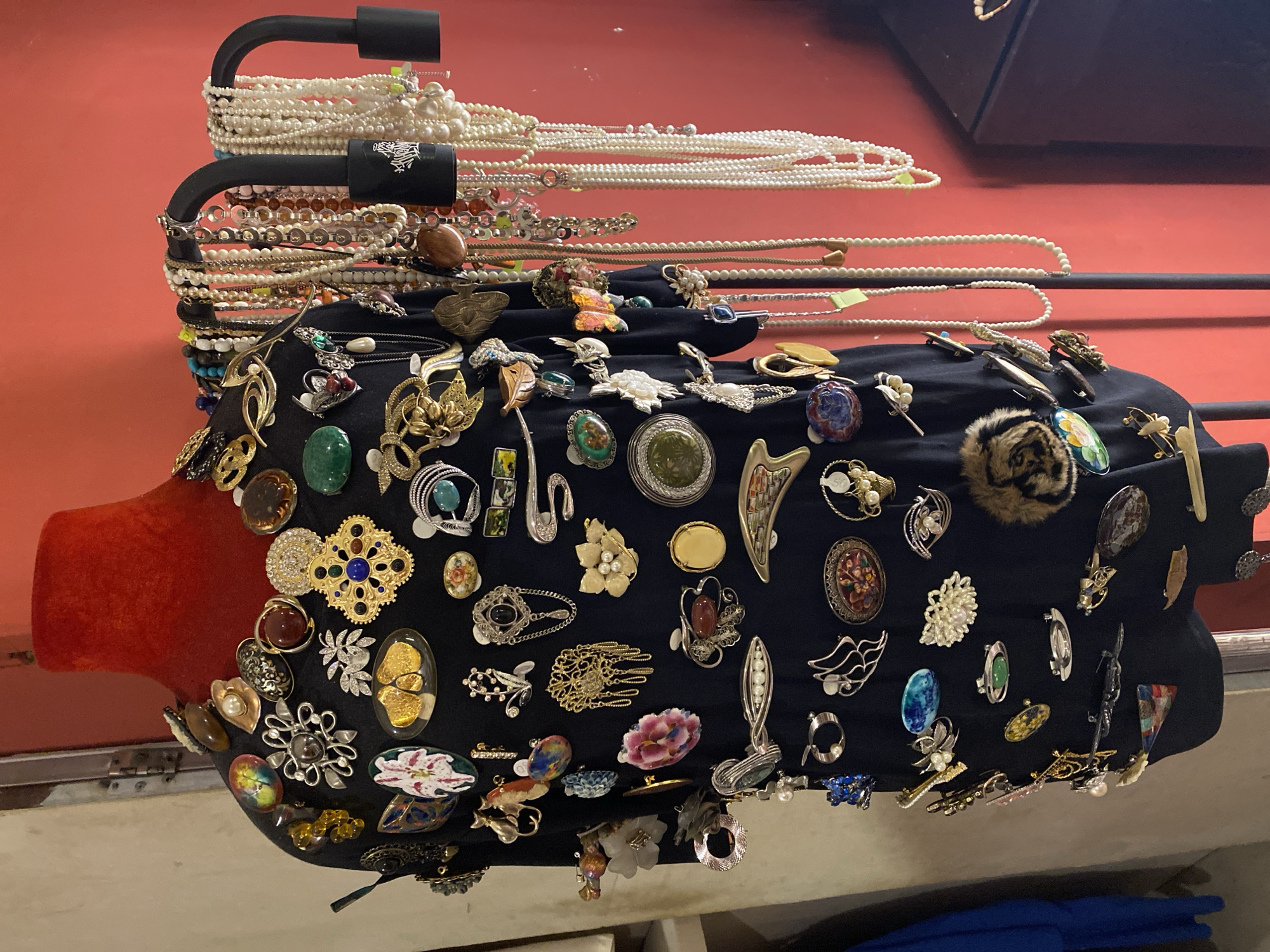 This image depicts a black garment, likely a shirt or dress, displayed on a red mannequin bust. The garment is heavily adorned with a large assortment of brooches and pins, creating a dense and varied pattern. The brooches are diverse in style, material, and design, including floral motifs, geometric shapes, animal figures, and abstract pieces. Some feature colored stones, pearls, and metalwork in gold, silver, and other finishes. The garment is positioned on a stand within what appears to be a retail or display space. To the right of the mannequin, a black metal rack holds numerous strands of necklaces, predominantly pearl and bead necklaces, of varying lengths. The background consists of a plain reddish-orange wall. The overall impression is of a display showcasing a collection of costume jewelry. There are no people visible in the image, and no specific location in Da Nang, Vietnam can be determined.