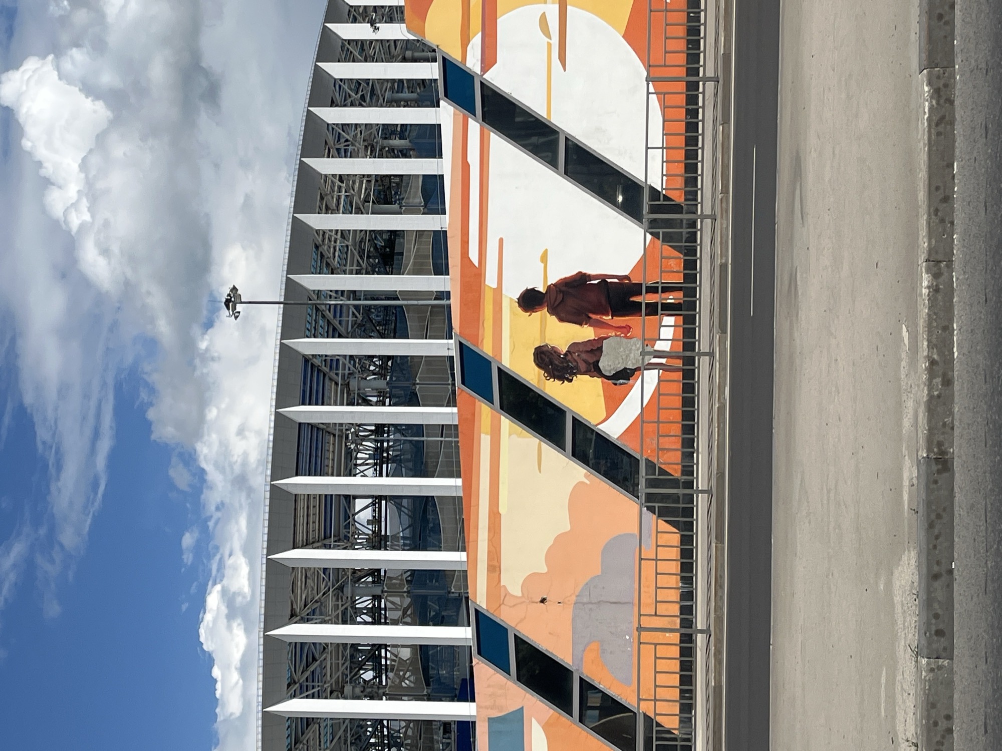 This image captures a mural on a wall in front of a large stadium structure. The mural depicts a young couple holding hands, rendered in an anime style, standing against a backdrop of stylized orange and yellow clouds and a setting sun. The stadium itself is a modern, multi-tiered structure with white vertical supports and a metallic roof, suggesting it might be a sports arena or concert venue. A street and sidewalk are visible in the foreground, with a metal fence separating the sidewalk from the mural. The sky above is blue with scattered white clouds, indicating it's a daytime scene.
This mural is located on the exterior wall of the Nizhny Novgorod Stadium, also known as "Nizhny Novgorod Arena," which was a venue for the 2018 FIFA World Cup.