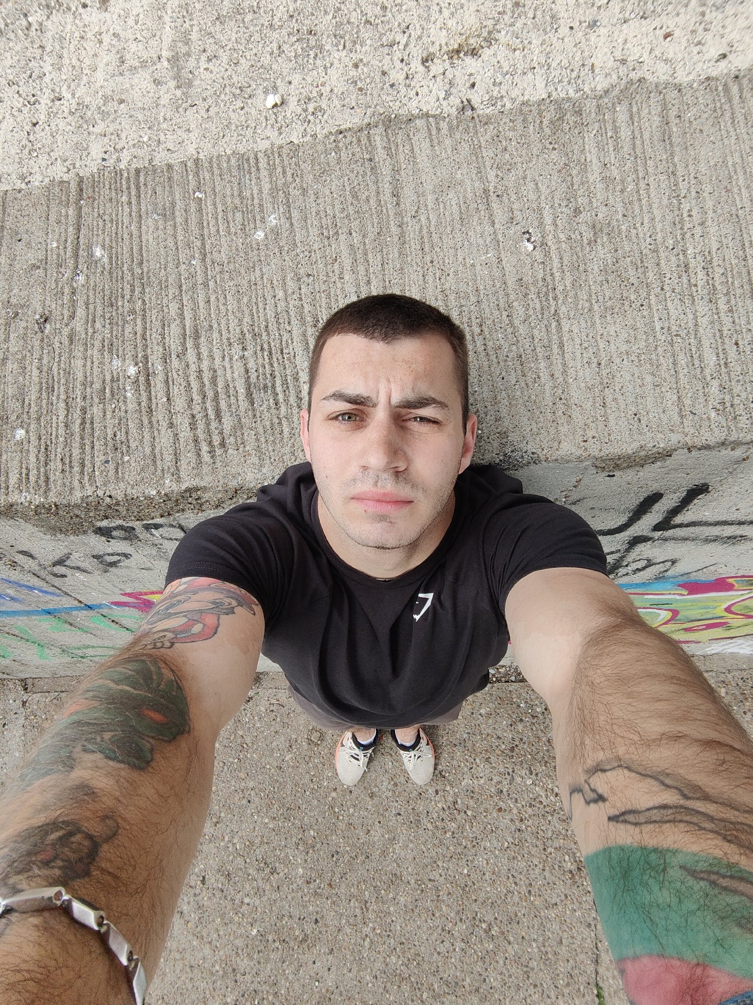 A man stands in front of a concrete retaining wall with graffiti on its lower portion. He is wearing a black t-shirt with a white logo, and his forearms are heavily tattooed. He also wears a silver bracelet on his left wrist. His feet, clad in white sneakers with blue laces, are visible near the bottom of the frame. The background consists of concrete pavement and the textured surface of the retaining wall. The photograph is taken from a high-angle perspective, suggesting the man is holding the camera above himself. The location is likely in Varna, Bulgaria, but there is no confirmation within the image.