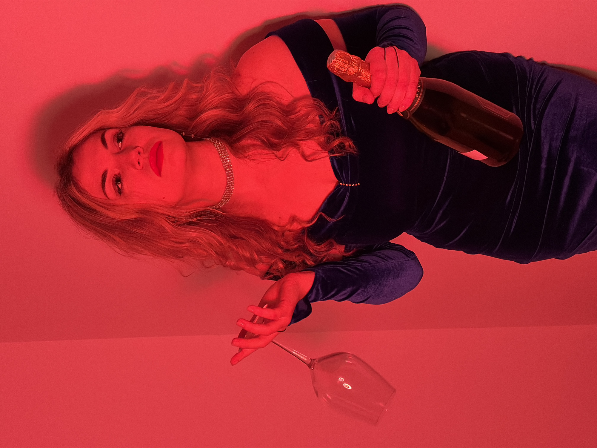 A woman lies on a red surface, wearing a blue dress. She holds a champagne bottle in her right hand and a wine glass in her left hand. The background is uniformly red. There is no visible indication of Budva, Montenegro.