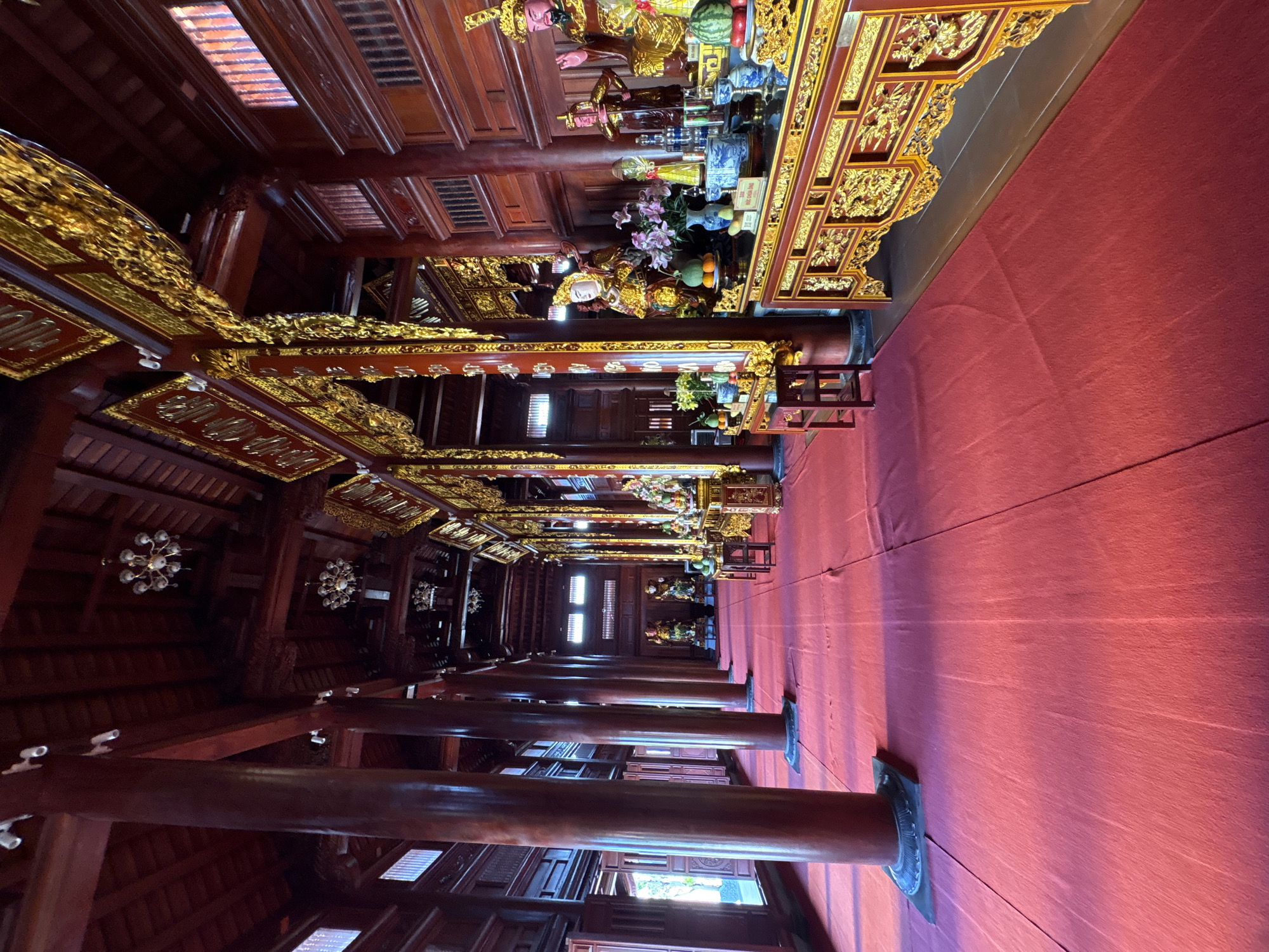 The image depicts the interior of a traditional Vietnamese building in Nam Nha Trang Ward, Vietnam. The space is defined by rows of polished wooden columns that support an ornate, gilded ceiling. At the top of the image, there is a red-carpeted area. The ceiling is decorated with intricate golden carvings. The overall atmosphere is one of formality and tradition.