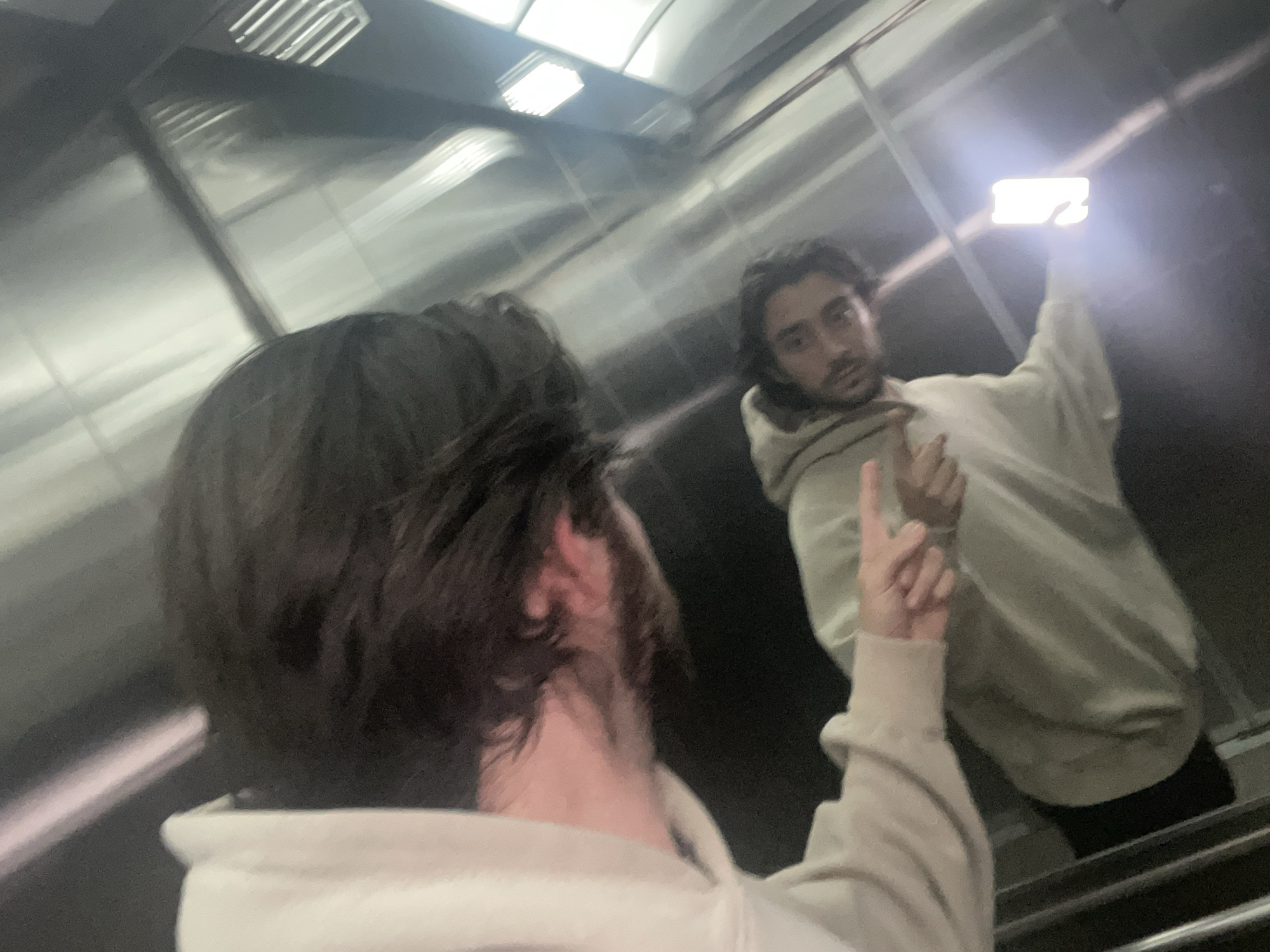 A man takes a selfie in an elevator. The interior of the elevator is metallic, with horizontal lines visible. The man taking the picture holds a glowing smartphone. The man is wearing a light-colored hoodie and black pants. A second person, also in a light-colored hoodie, is partially visible in the foreground. The exact location is not discernible from this image and it cannot be confirmed that the picture was taken in Huixquilucan de Degollado, Mexico.