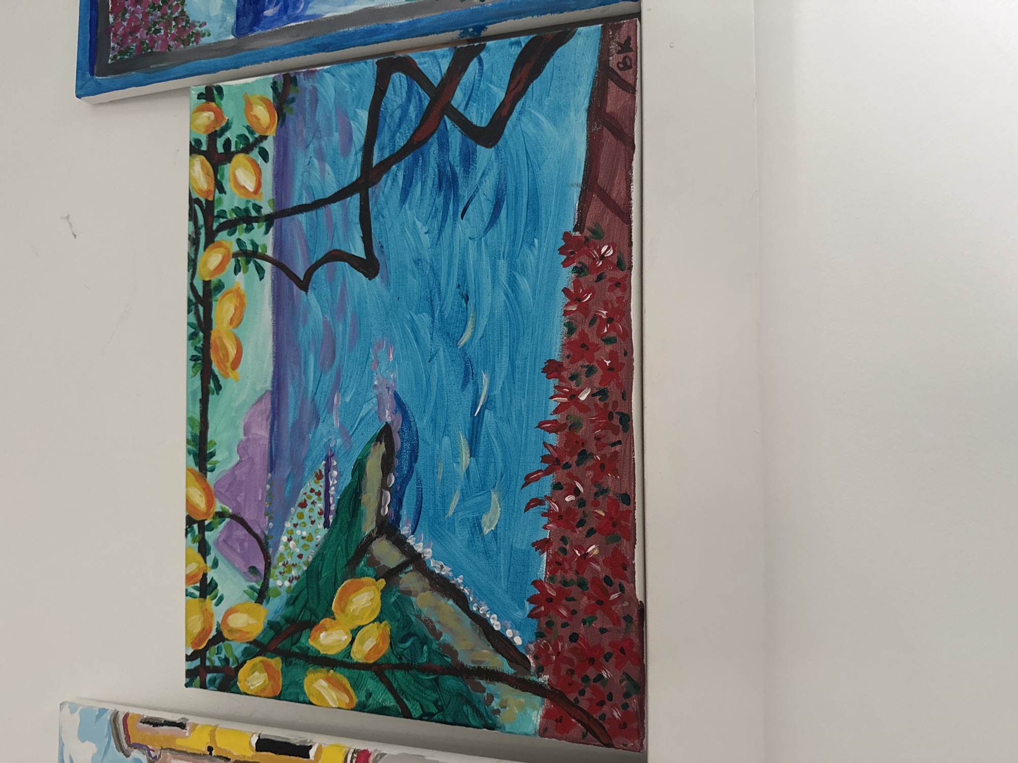 The image shows a painting hung on a white wall. The painting depicts a scene with various elements including yellow fruits (possibly lemons), red flowers, and a blue background that suggests a sky or water. The artistic style is somewhat naive or impressionistic. The painting is hung vertically on the wall, with other artwork visible above and below. The environment appears to be an indoor space, possibly a gallery or a home. There is no indication of a specific location or city, and no people are visible. The only objects present are the painting and the wall.