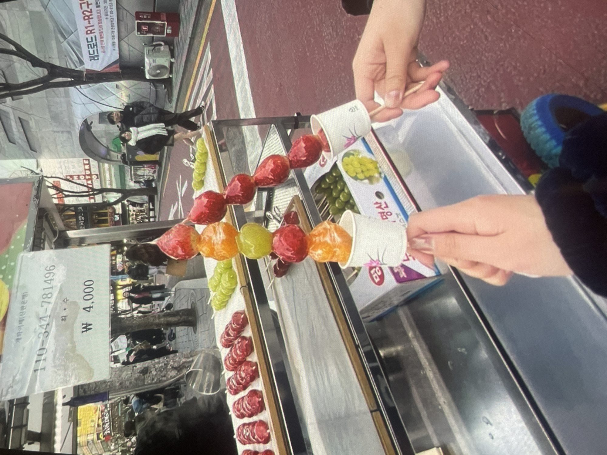 The image captures a street food scene, likely in South Korea, as indicated by the Korean language on the price sign (₩ 4,000) and the presence of Korean street food. The main subject is a vendor selling fruit skewers, called Tanghulu, glazed with a sugary coating. Various types of fruit, including strawberries, grapes, and mandarin oranges, are visible on the skewers. Two hands are holding the skewers in white paper cups. The price sign is prominently displayed, showing a bank account for transfer payments. In the background, a street scene with pedestrians and shops is visible. There is no indication that the image is related to the city of Harmanli, Bulgaria.