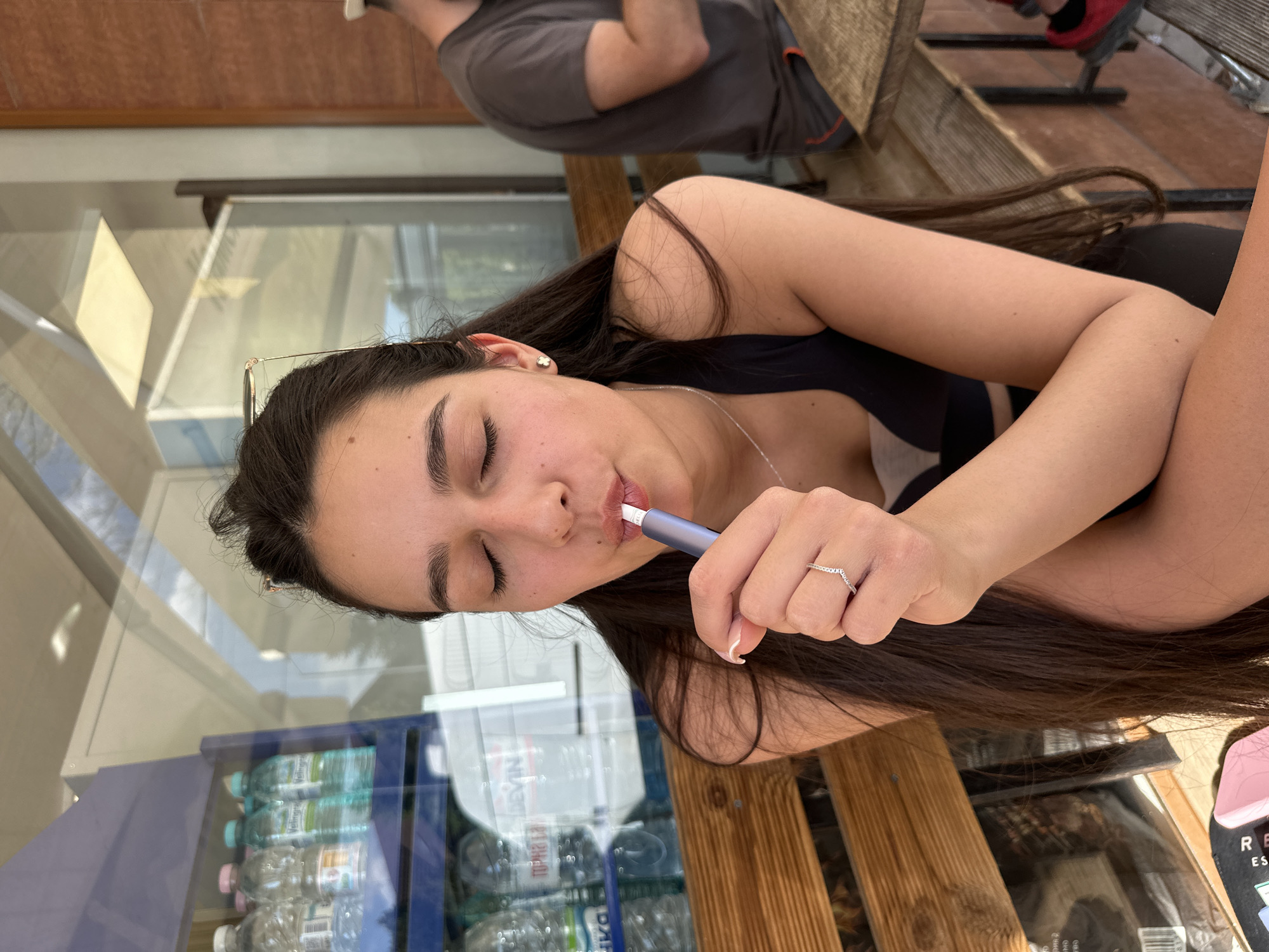 The image shows a young woman sitting at an outdoor table. She is wearing a black top and has long, dark brown hair. She is holding a blue and white electronic cigarette to her lips with her eyes closed. She is wearing a silver ring on her ring finger. Behind her is a glass-fronted cooler filled with bottles of water. Wooden beams form the structure of the outdoor seating area. Another person is partially visible behind her wearing a grey shirt. The location is likely an outdoor café in Plovdiv, Bulgaria, given the presence of Cyrillic script on the water bottle labels.