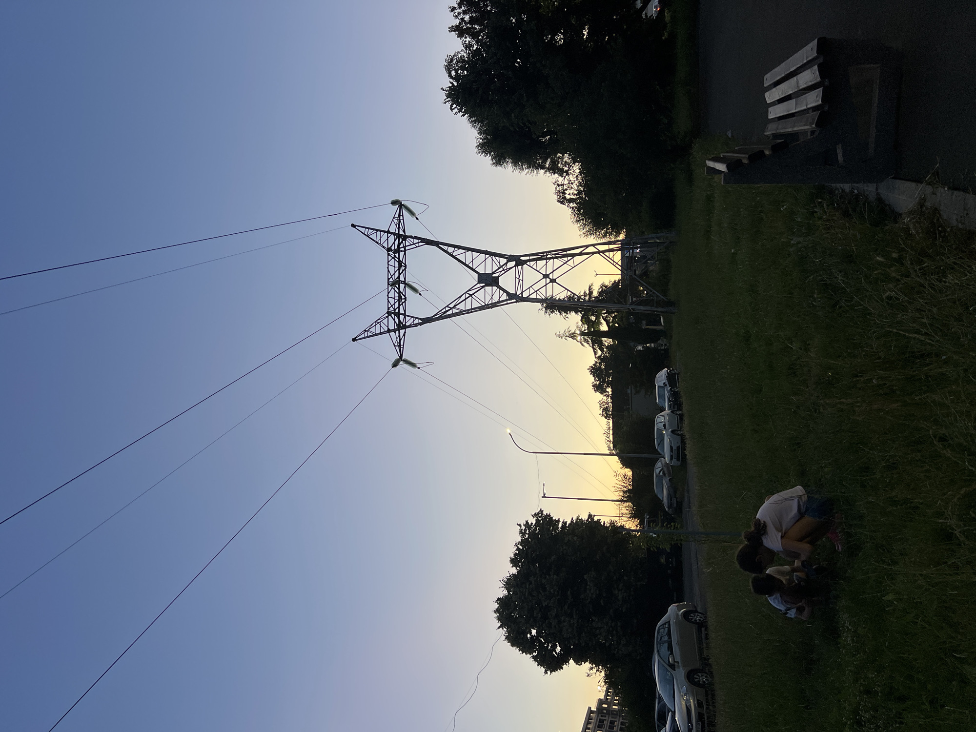 The image depicts a park scene in the city of Burgas, Bulgaria, during twilight. Dominating the background is a tall, industrial-looking electricity pylon with power lines extending upwards and across the sky. The sky transitions from a pale blue at the top to a warm yellow and orange hue near the horizon, suggesting sunset or sunrise.
In the foreground, two individuals, an adult and a child, are crouched down in the grass, seemingly engaged in an activity together. The adult, wearing a light-colored t-shirt and shorts, is leaning over. The child, wearing a darker outfit, is positioned close to the adult. They are surrounded by green grass and some weeds.
To the left of the scene, cars are parked along what appears to be a road or parking lot, with trees and buildings visible further in the background. To the right, a park bench made of wood and stone sits on a paved area next to the grass. Trees with dark foliage are scattered throughout the middle and right side of the frame. Streetlights are also visible, casting a soft glow. The overall atmosphere is calm and serene, transitioning from daylight to evening.