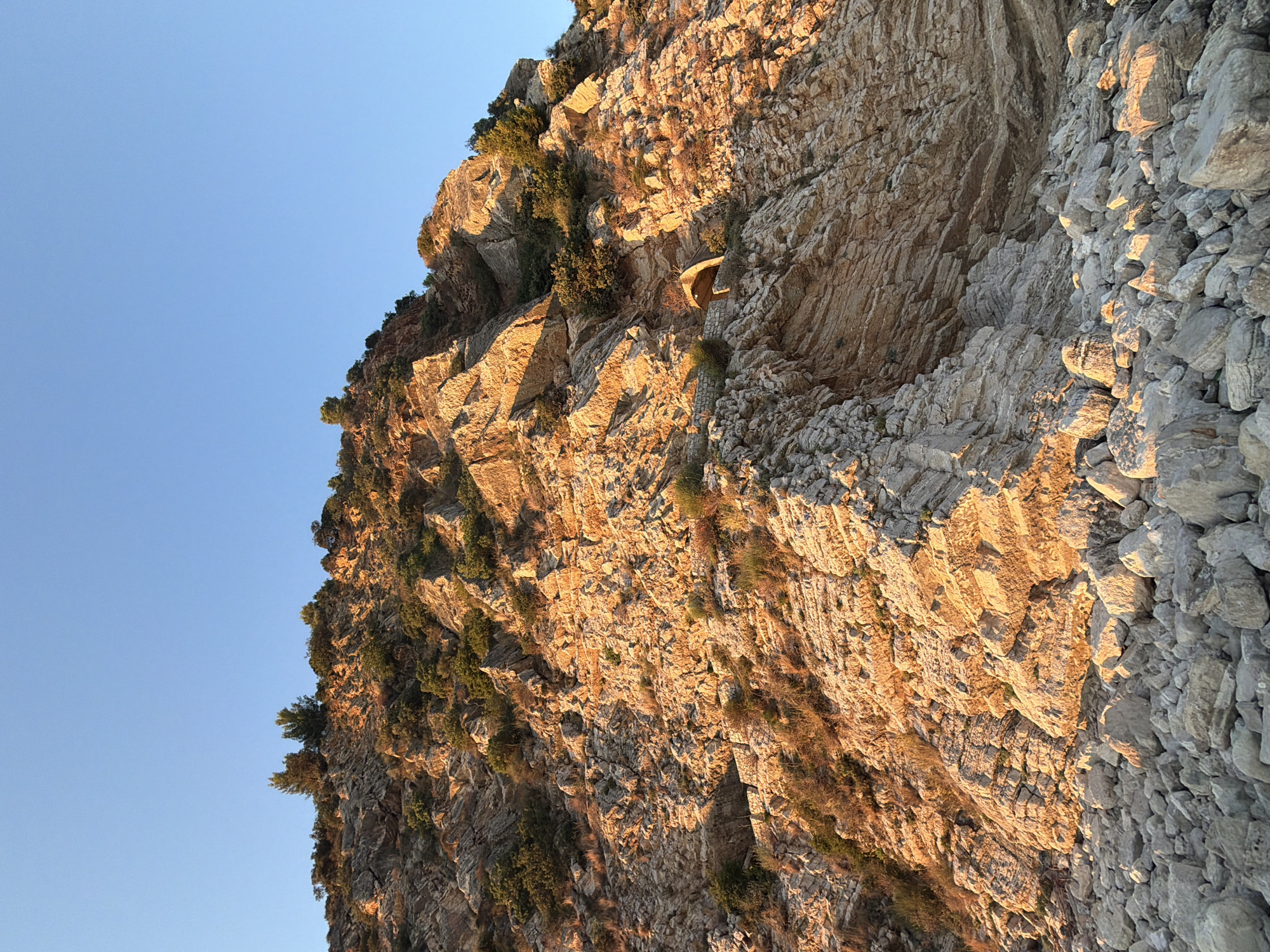 A low-angle, outdoor, full shot captures a rocky mountainside in Novoselje, Montenegro, under a clear, pale blue sky during what appears to be sunset. The mountain, illuminated by warm, golden sunlight, is characterized by jagged rock formations with distinct horizontal striations. Patches of green foliage and dry, brown vegetation are scattered across the slopes. The foreground is filled with a dense collection of large, gray rocks. In the middle of the mountain face, a natural archway or cave entrance is visible, partially obscured by shadows and vegetation. No people or animals are present in the scene.