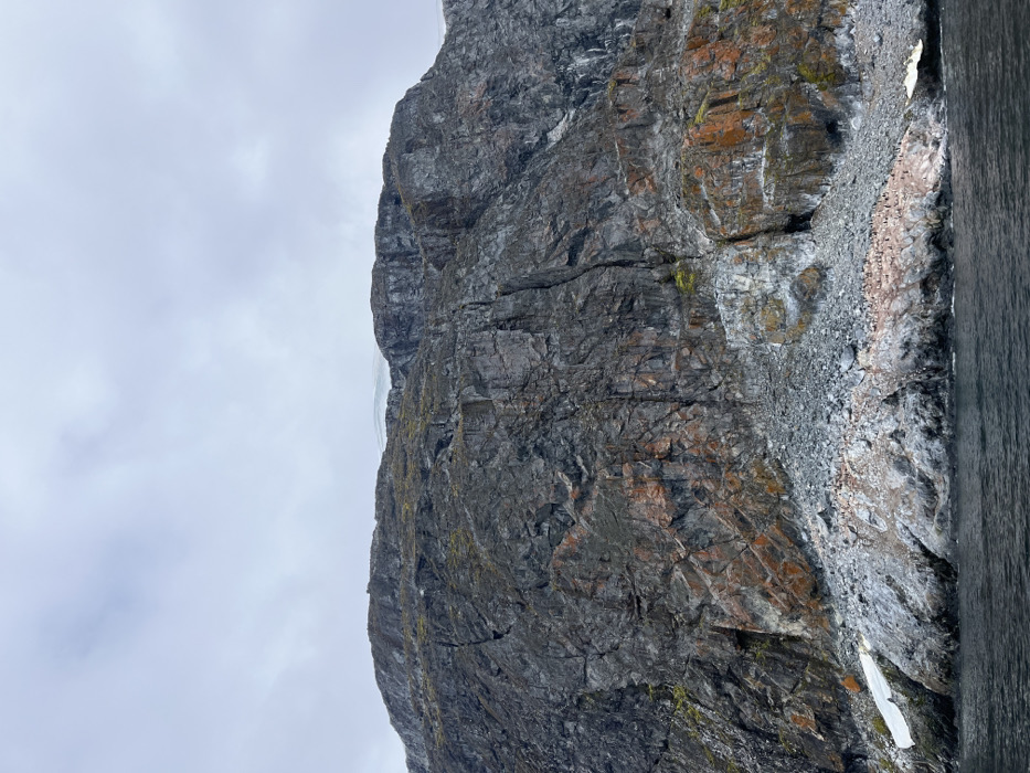 A towering, rocky cliff face dominates the frame, its surface etched with deep fissures and variations in color from dark gray to rusty orange. Sparse patches of green moss cling to the rock. A cloudy sky occupies the left portion. On the right, a dark, smooth surface of water or another dark object is barely visible, with some loose stones and snow near the cliff base. The image is oriented vertically.