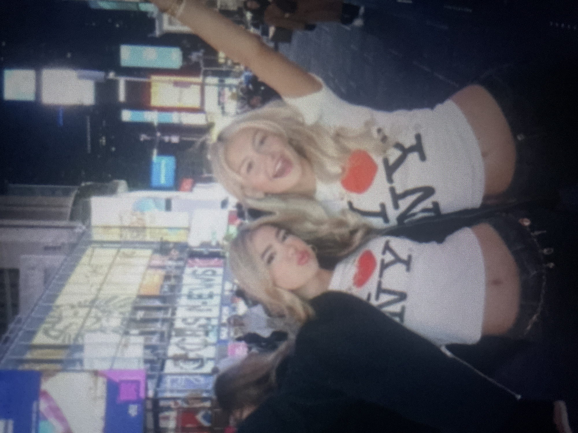 Two young women are posed in Times Square, New York City, at night. Both are wearing white crop tops with "I ❤️ NY" printed on them. The woman on the left has her hair down and is puckering her lips. The woman on the right has her hair down and is smiling widely with her mouth open, raising her right arm in the air. The background shows the bright lights and billboards of Times Square, with various buildings and people visible.