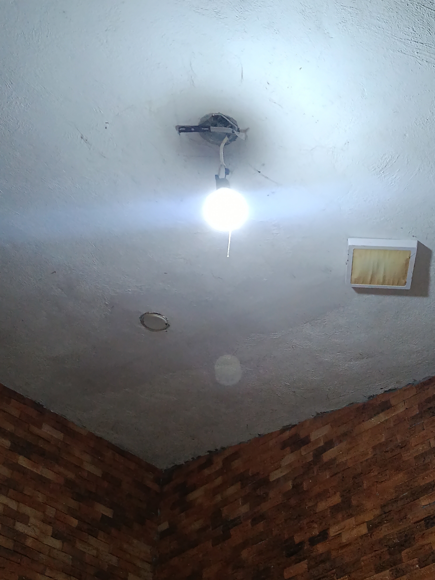 The image shows a corner of a room with a brick wall and a white textured ceiling. A bright light bulb is hanging from the ceiling, illuminating the area. To the right of the light bulb, there is a rectangular white object with a yellowed panel, possibly an air vent or a light fixture. There is also a circular fixture embedded in the ceiling further to the left. The brick wall appears to be made of reddish-brown bricks laid in a stretcher bond pattern. The lighting in the image suggests it may be indoors, possibly in a residential or commercial space in Juan Griego, Venezuela, given the context. There are no people or explicit actions depicted in the image.
