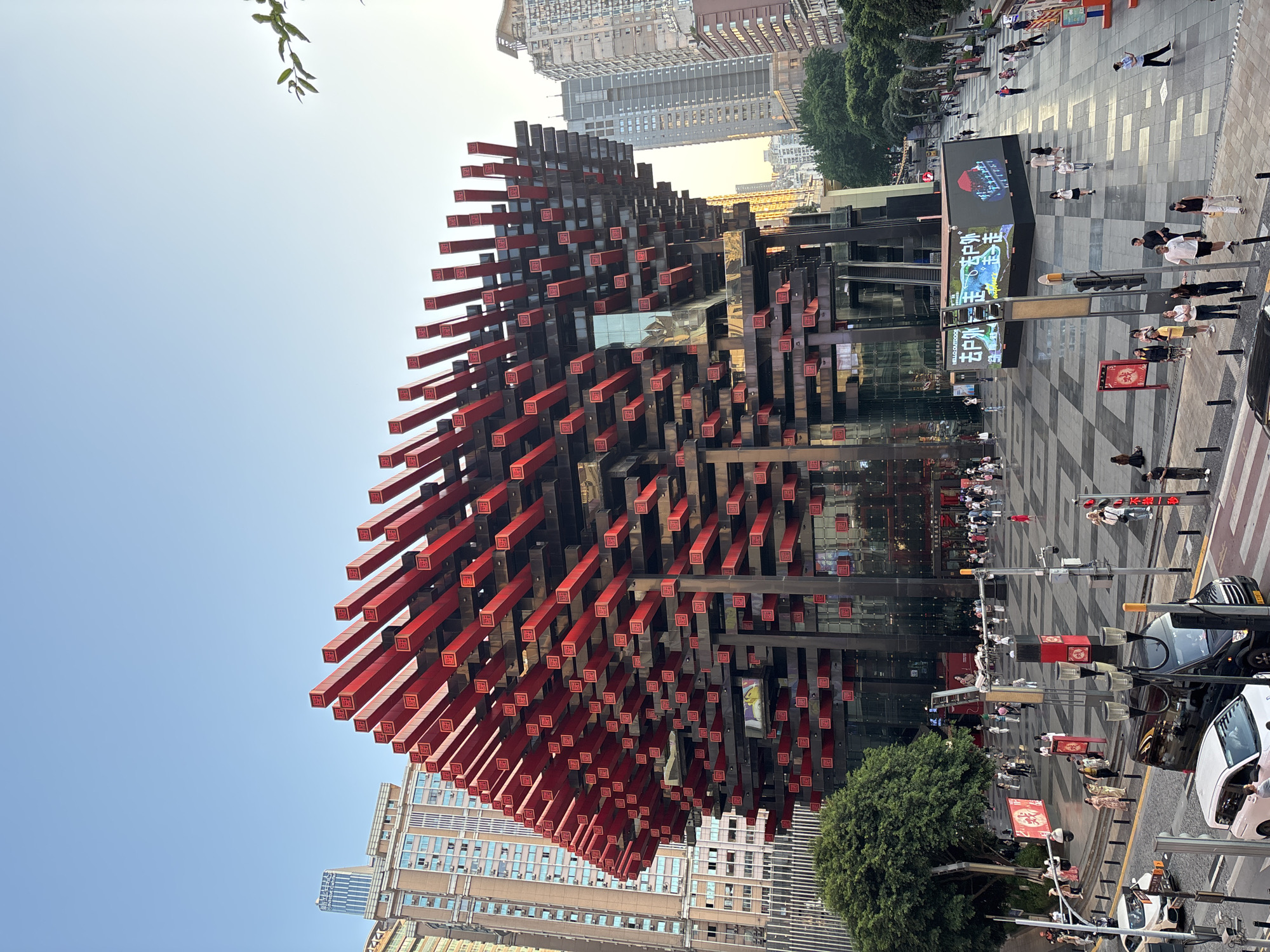 The image depicts the Raffles City Chongqing complex in Yuzhong District, China. The foreground features a pedestrian area with people walking, including groups and individuals. The building's distinctive facade is visible, characterized by red and black rectangular extrusions. Trees are seen alongside the road, and cars are parked on the street. A large screen displays signage in Chinese. The background shows other tall buildings, typical of an urban environment. The sky is light blue, indicating daytime.