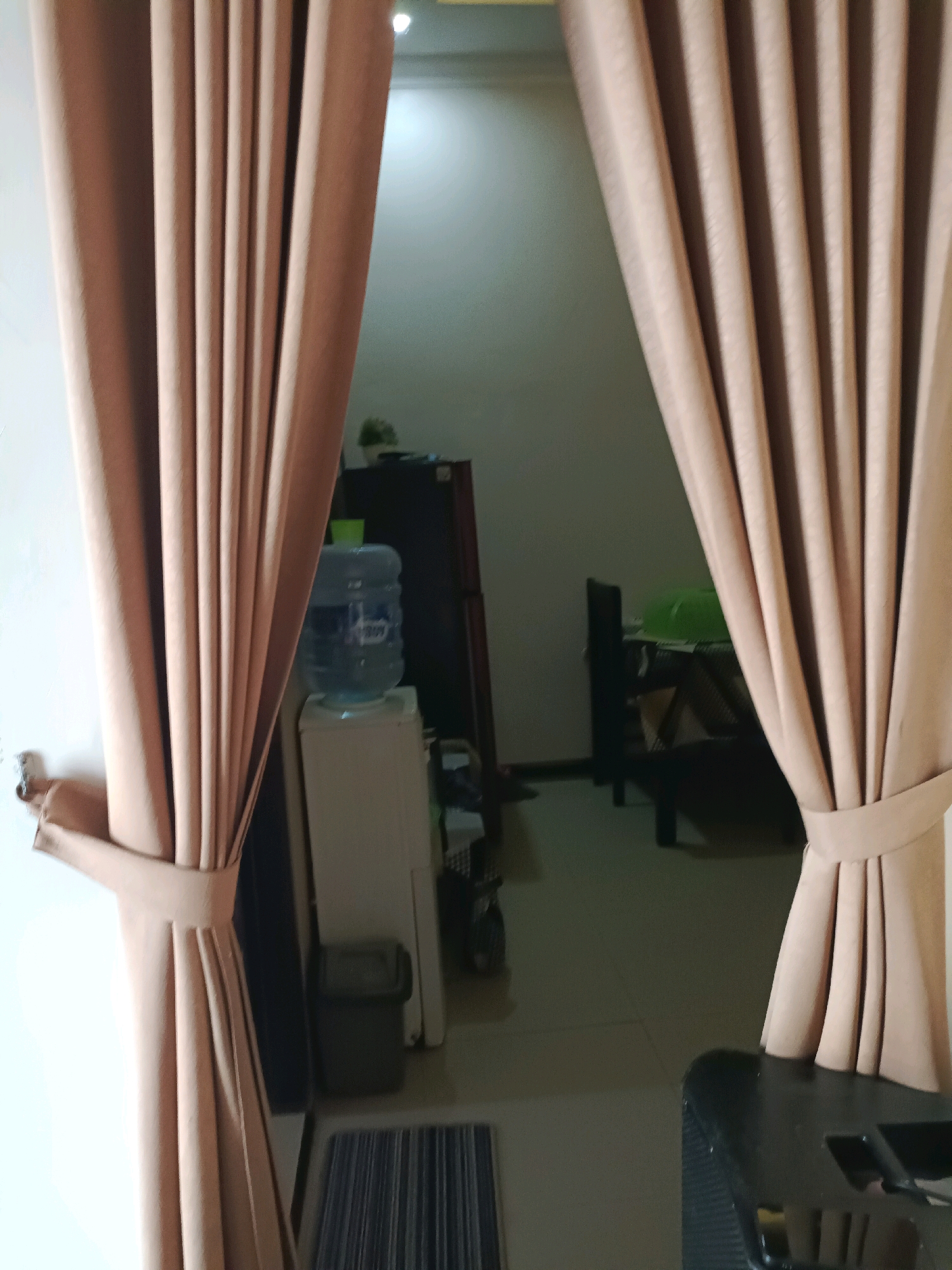 In the city of Rancang Kencono, Indonesia, an interior scene is depicted, dominated by the presence of light brown curtains that frame the view into another room. The curtains are gathered and tied back with matching fabric bands. To the left, a white wall is visible, with a small piece of hardware attached.
Through the gap in the curtains, a white water cooler with a large blue water jug on top is present. Adjacent to the water cooler is a dark refrigerator. In the background, a table with dark legs and a light-colored surface is partially visible, with a green object on it. The floor appears to be a dark, reflective surface. A striped mat is positioned on the floor in the foreground, to the right of the water cooler. A black object with a textured surface is in the lower right corner of the image. The overall lighting suggests an indoor environment with ambient light coming from the room beyond the curtains.