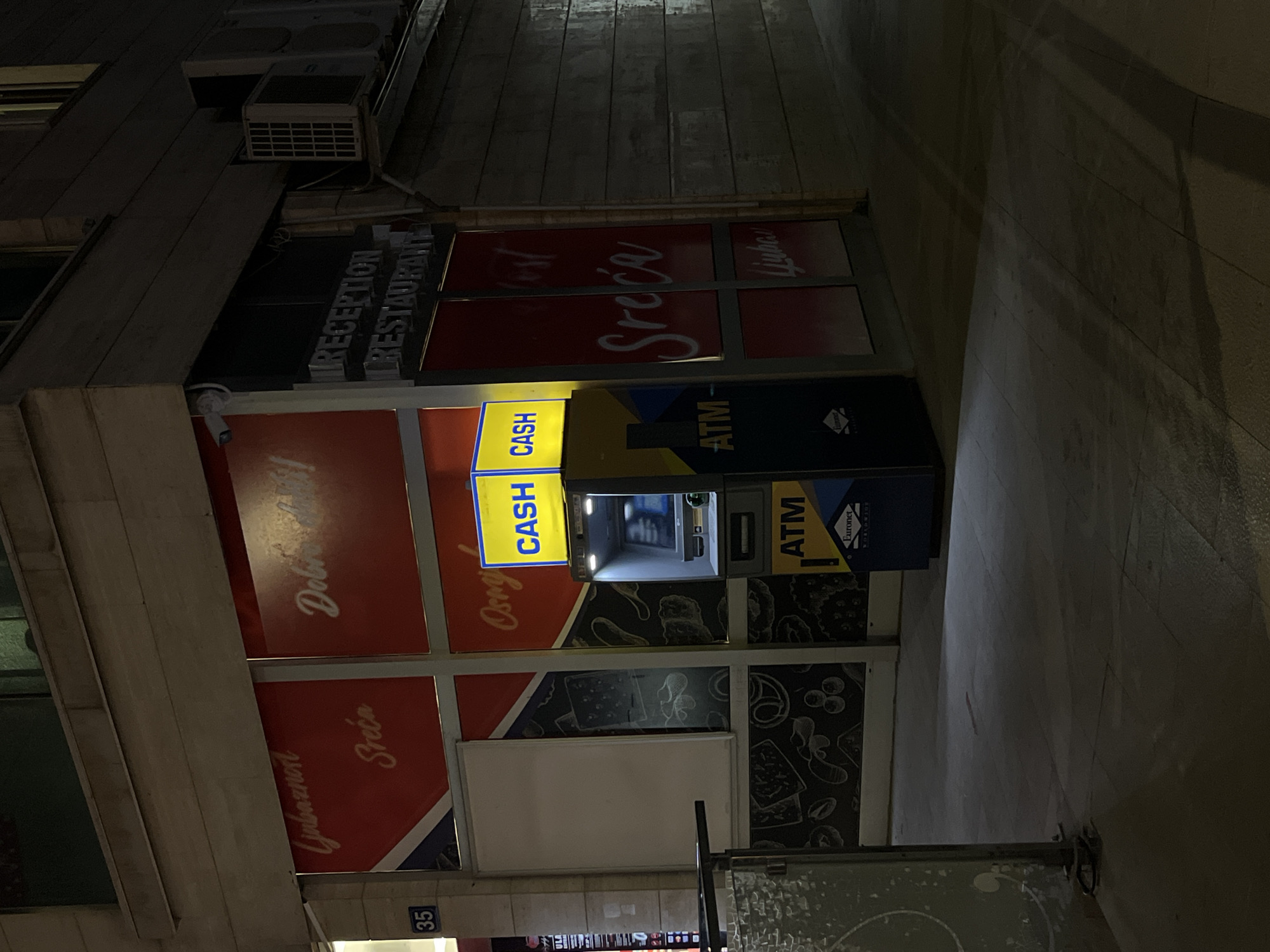 The image depicts an exterior view of a building at night, likely in Budva, Montenegro, featuring an ATM and storefronts. The ATM, a prominent yellow and blue machine, is illuminated by a bright yellow sign displaying "CASH" in blue letters. Below the "CASH" sign, the word "ATM" is visible in white on a blue background. The ATM itself is a silver-colored machine with a screen and keypad.
To the left of the ATM, the storefront has red panels with white lettering that appears to be in a Slavic language, possibly reading "Ljubaznost" and "Sreca," which translate to "Kindness" and "Luck" respectively in English. Further to the left, a blue sign with the number "35" is affixed to a concrete pillar.
To the right of the ATM, a section of the building has a sign reading "RECEPTION RESTAURANT" in silver lettering. This area also has a white outdoor air conditioning unit mounted on the wall. The ground in front of the building is paved with stone tiles, and shadows are cast across it due to the nighttime lighting. A metal barrier with a decorative pattern is visible in the lower left corner of the image. The overall impression is a well-lit commercial facade in an urban setting during the evening.