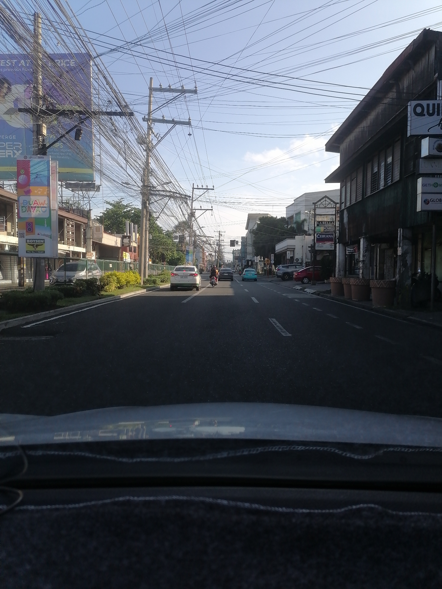 The image presents a forward-facing perspective from inside a vehicle traveling along an urban street in Davao City, Philippines. The sky above is clear and bright blue, indicating fair weather. The roadway is paved with asphalt, featuring white dashed lane markings.
Overhead, an intricate and dense network of black utility wires crisscrosses between numerous wooden and concrete utility poles lining both sides of the street.
On the left side of the road, a concrete median is adorned with green and yellow shrubs. Behind this median, a large billboard is prominently displayed, featuring the text "DUAW DAVAO" and "BOYSEN The No. 1 Paint," alongside other advertising elements like "BEST AT PRICE" and "320 mbps." Further back, a white van is parked near commercial buildings.
On the right side of the road, an old, two-story building with a brown roof and a dark green or grey facade stands out. Signs attached to this building include partial lettering for "QUI," "GETHOM," and "GLOBAL." A line of large, terracotta-colored plant pots sits along the building's frontage. Further down the street on the right, other businesses are visible, identified by signs such as "LA BELLA" and "CEBUANA."
Traffic on the road includes a white sedan with license plate JAF 3395 and a motorcycle following it in the left lane. Other vehicles, including a red car and a light blue sedan, are visible further down the road. The lower portion of the image displays the dark dashboard and a slight reflection from the vehicle's windshield.