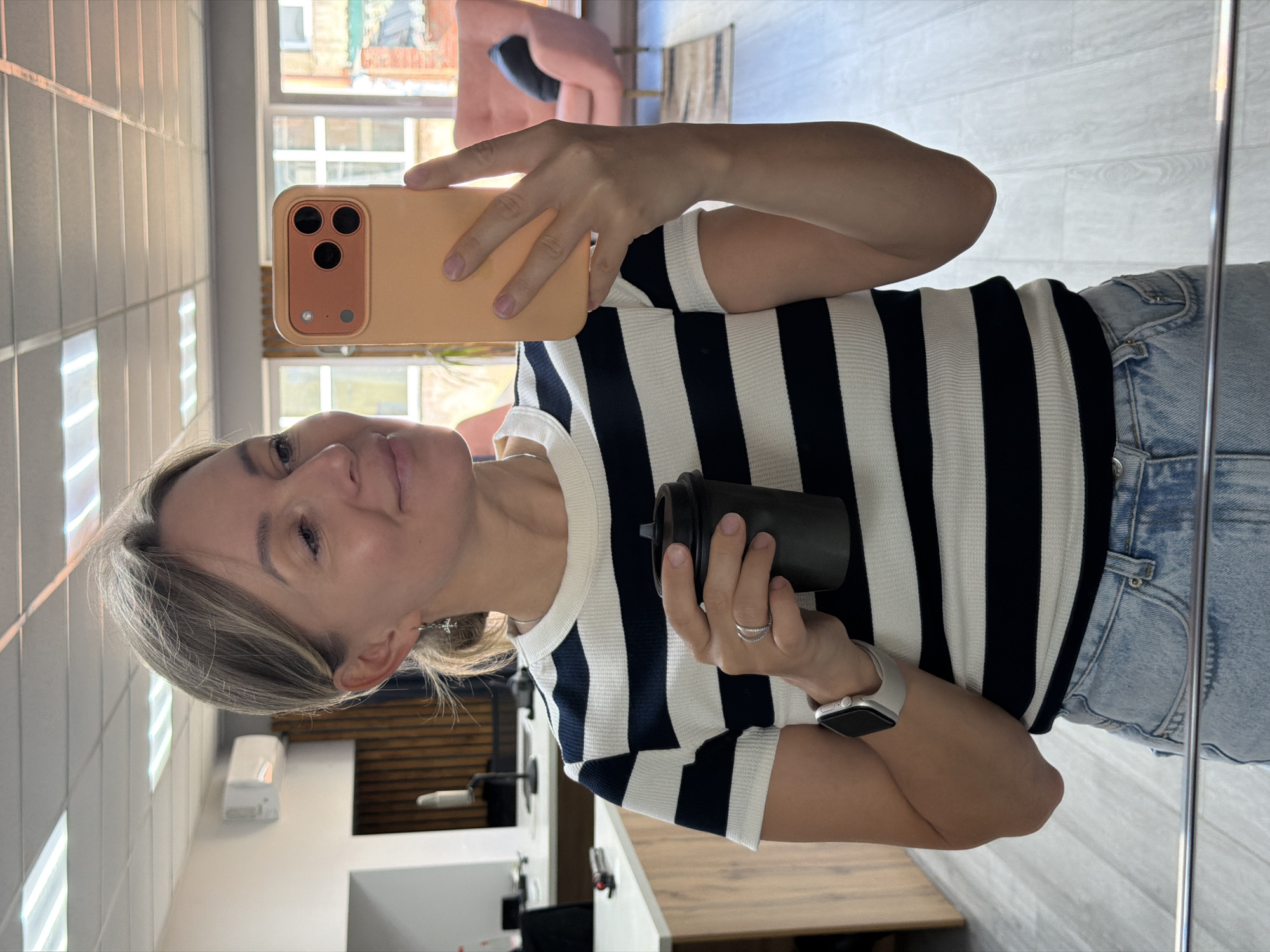 A woman is taking a mirror selfie in what appears to be a salon or beauty studio. She is holding an orange phone in her right hand, using it to capture her reflection. She's wearing a black and white striped long-sleeved shirt and light blue denim jeans. On her left wrist, she has a silver smartwatch. She is also holding a black cylindrical object, possibly a hair product or a drink, in her left hand. Her blonde hair is pulled back, and she is wearing subtle makeup. She has a slight smile and seems relaxed. The background shows a tiled wall, fluorescent lighting, and a window with sunlight streaming in, suggesting it's daytime. In the reflection, another part of the room is visible, including a pink chair and some wooden paneling. There is no visible text in the image. The context provided indicates the location is Chaikovsky, Russia.