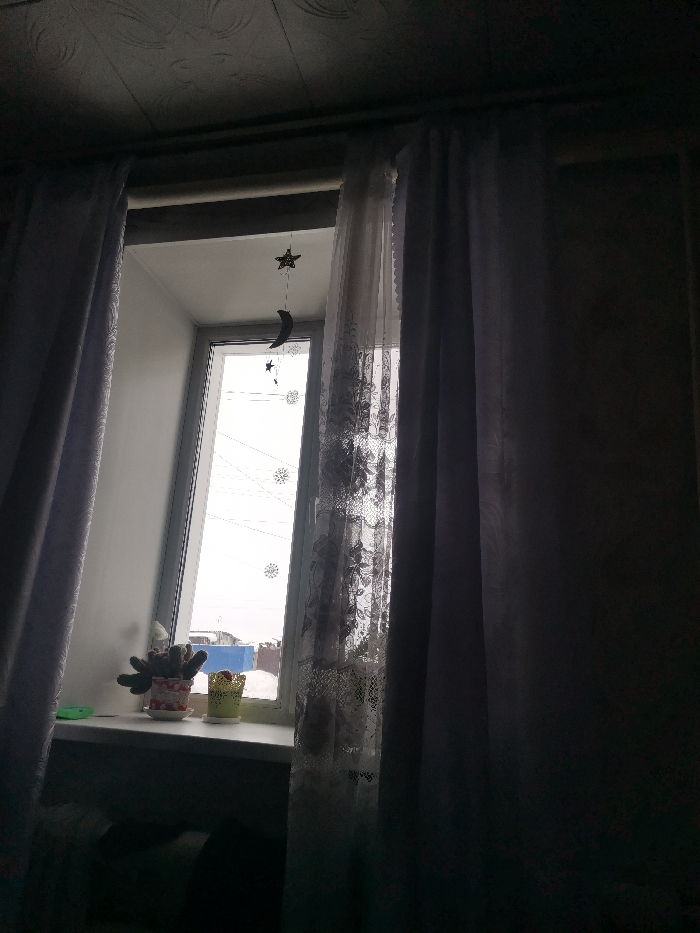 A window partially obscured by grey and white curtains. A star and moon mobile hangs in front of the glass. Two potted plants sit on the windowsill. Outside, snow and houses are visible. The room is dark, illuminated only by the light filtering through the window. The overall tone is melancholic.