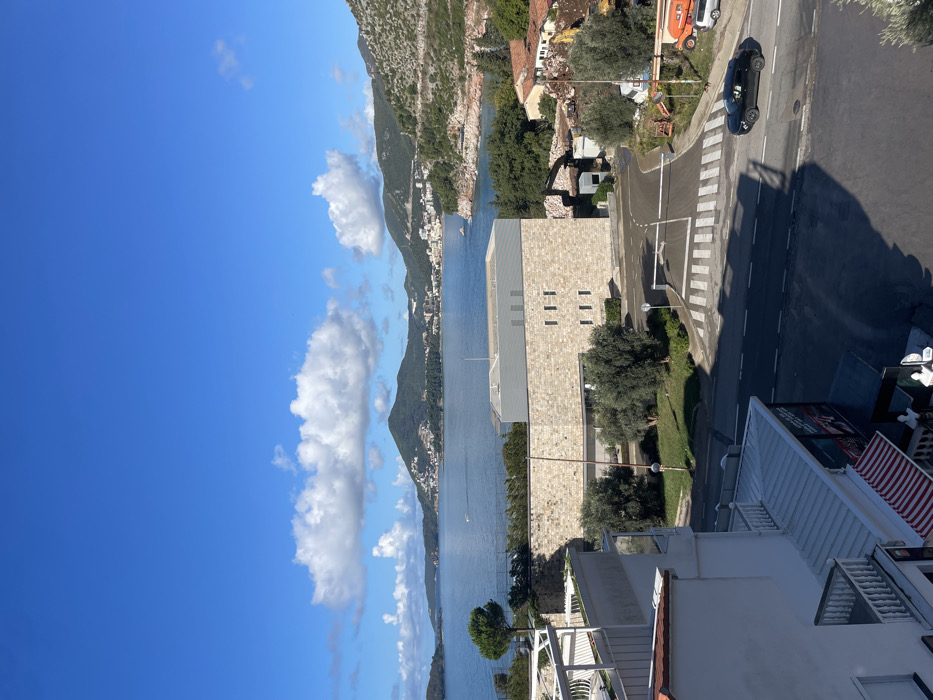 The image displays an elevated view of a coastal area within the city of Vrijesno, Montenegro, under a bright blue sky dotted with scattered white cumulus clouds. In the background, a large expanse of deep blue water, presumably a bay, extends towards a distant coastline characterized by rolling hills and mountains densely populated with residential and commercial buildings. Several small watercraft are visible on the bay.
In the mid-ground, a prominent modern building constructed of light-colored, textured stone cladding features multiple small, evenly spaced rectangular window openings. Adjacent to this structure on the right, a demolition or construction site is apparent, with an excavator and an orange concrete pump or mixer visible among debris and several smaller, traditional buildings with red-tiled roofs. A paved road with a white pedestrian crossing stripe and a horizontal white barrier arm traverses the scene. Mature green trees and various other vegetation are present around these structures and along the road.
In the immediate foreground, the top and side of a white multi-story building are partially visible on the left, featuring multiple balconies with white railings and a ground-level red and white striped awning. A dark-colored SUV is observed in motion on the road, traveling from right to left. The scene is brightly lit by direct sunlight, casting distinct shadows.
