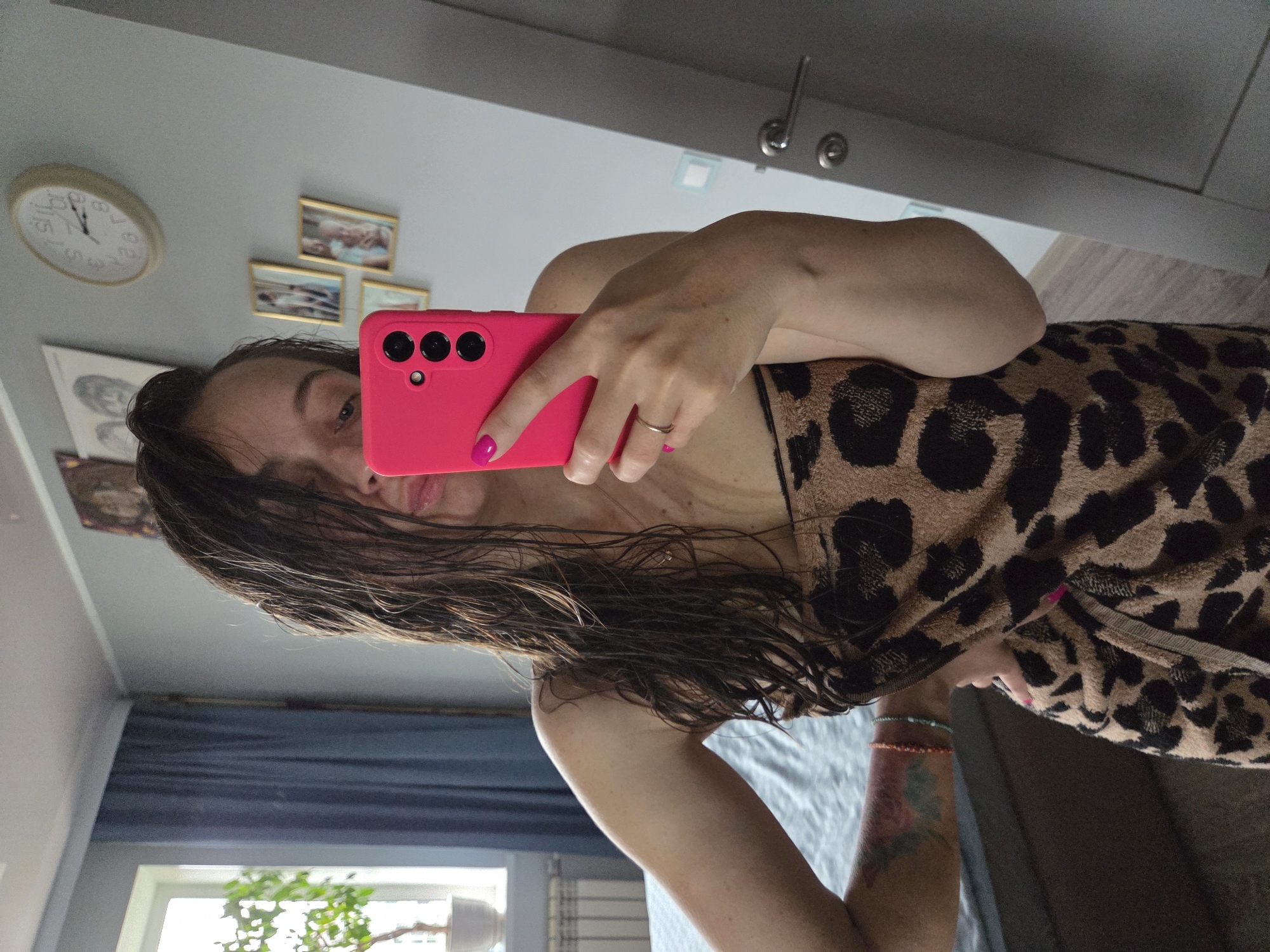A woman is taking a selfie in what appears to be a bedroom. She holds a pink smartphone with three camera lenses in her right hand. The woman has wet, dark hair and is wearing a towel with a leopard print design. A gold ring is visible on her finger. The room contains various elements: a clock and framed pictures on the wall, a window with a plant in front of it, and a door with a handle. The location of the city of Birobidzhan, Russia, cannot be determined from the given image.
